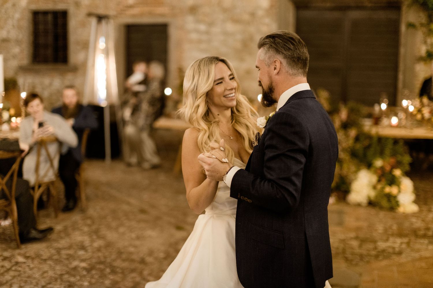 A couple shares a romantic moment during their evening wedding celebration at a rustic stone venue with glowing lights.