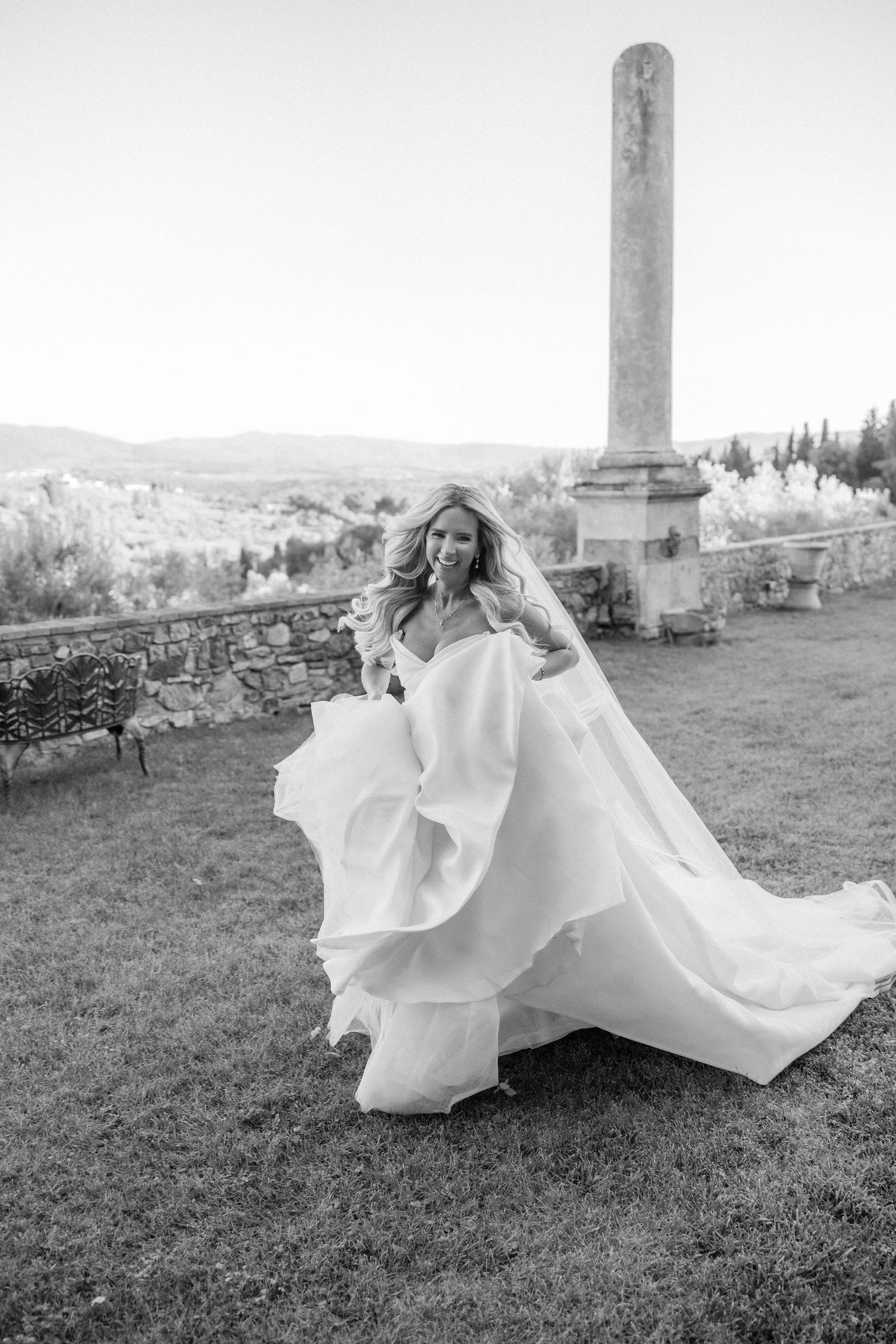 A flowing white wedding dress billows dramatically across a grassy hilltop setting in black and white.