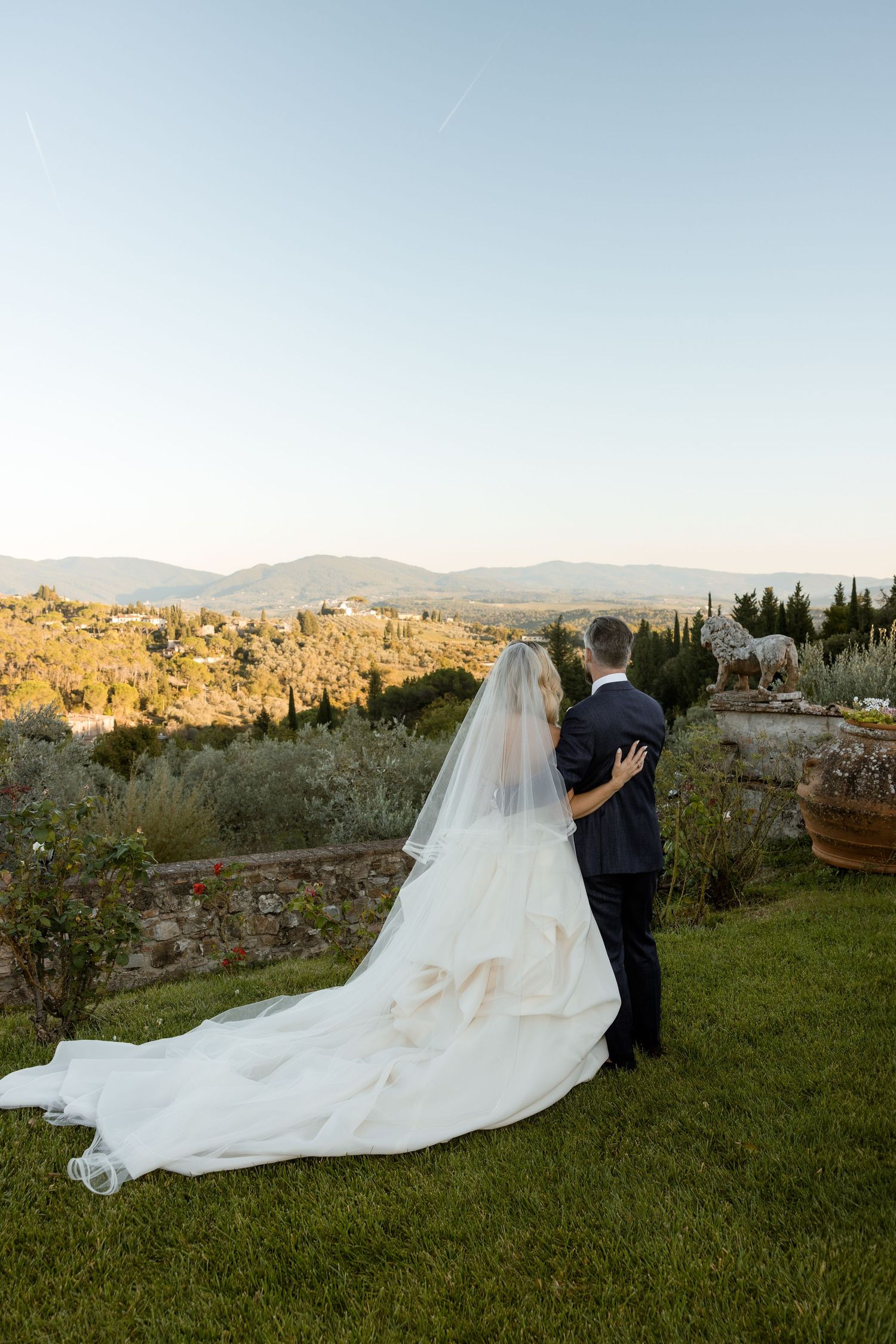 A bride and groom share a romantic moment on a hilltop overlooking a scenic Tuscan landscape at sunset.