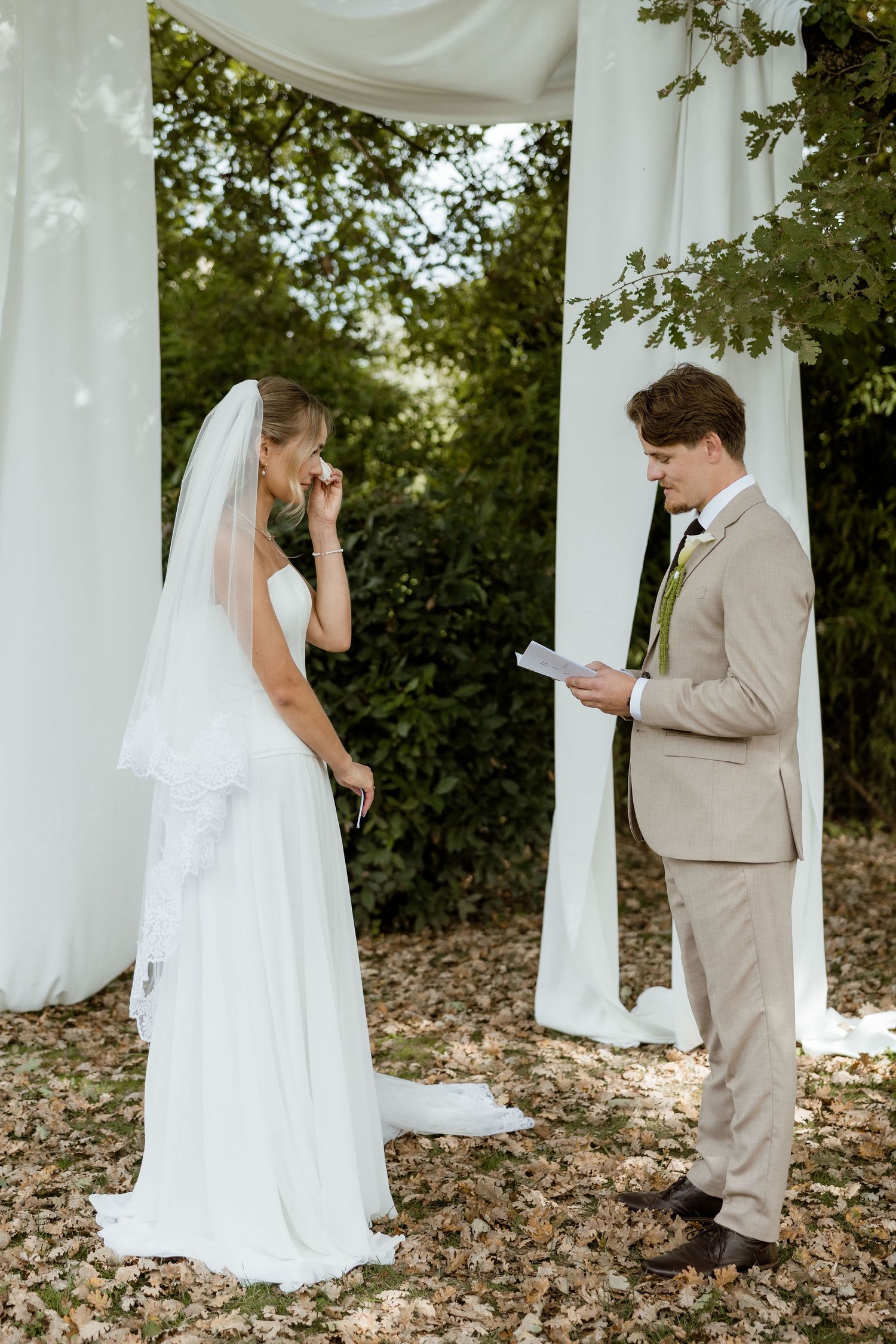 A couple in wedding attire share a private first look moment behind white curtains in a wooded outdoor setting.