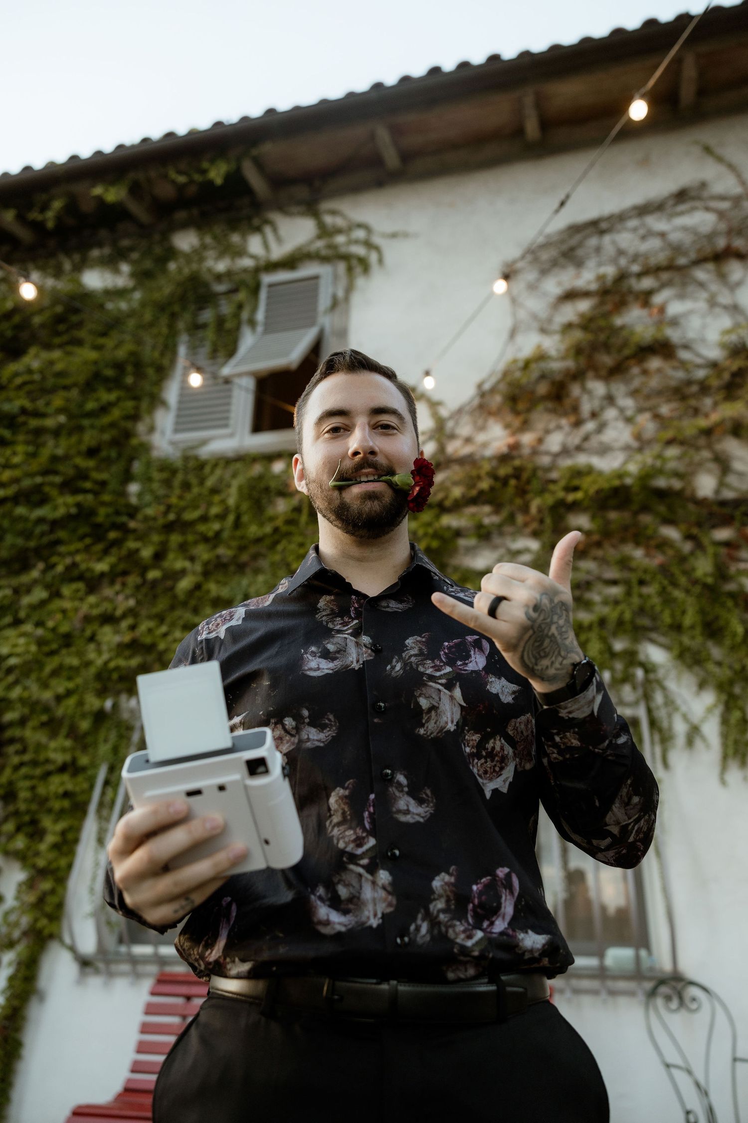 Person in black patterned hoodie holding and gesturing with a gaming device against white wall and string lights.