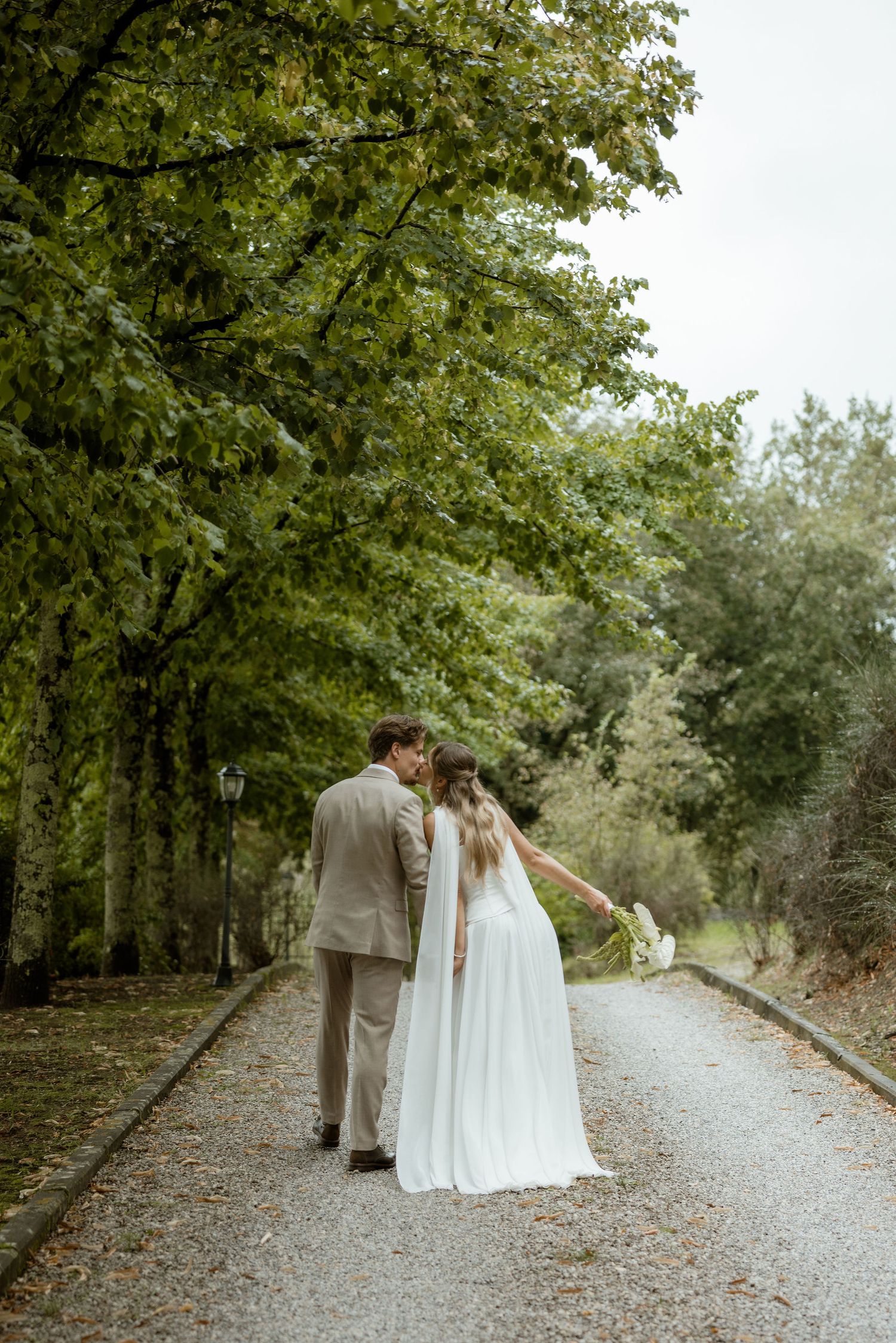 A romantic wedding couple walks together down a scenic tree-lined path during an autumn day.