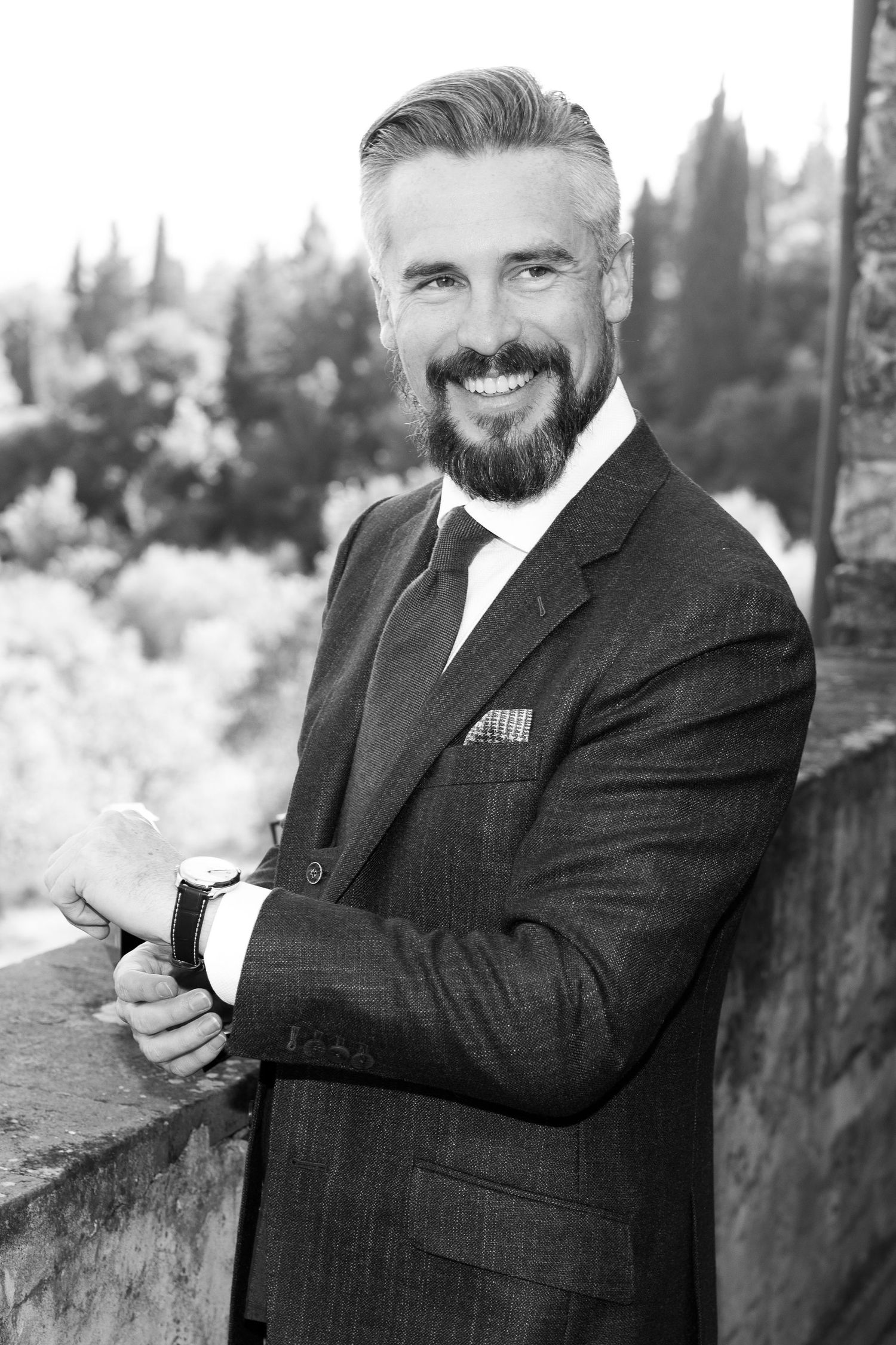 Professional portrait of a bearded man in a dark suit smiling and posing against a black and white background.