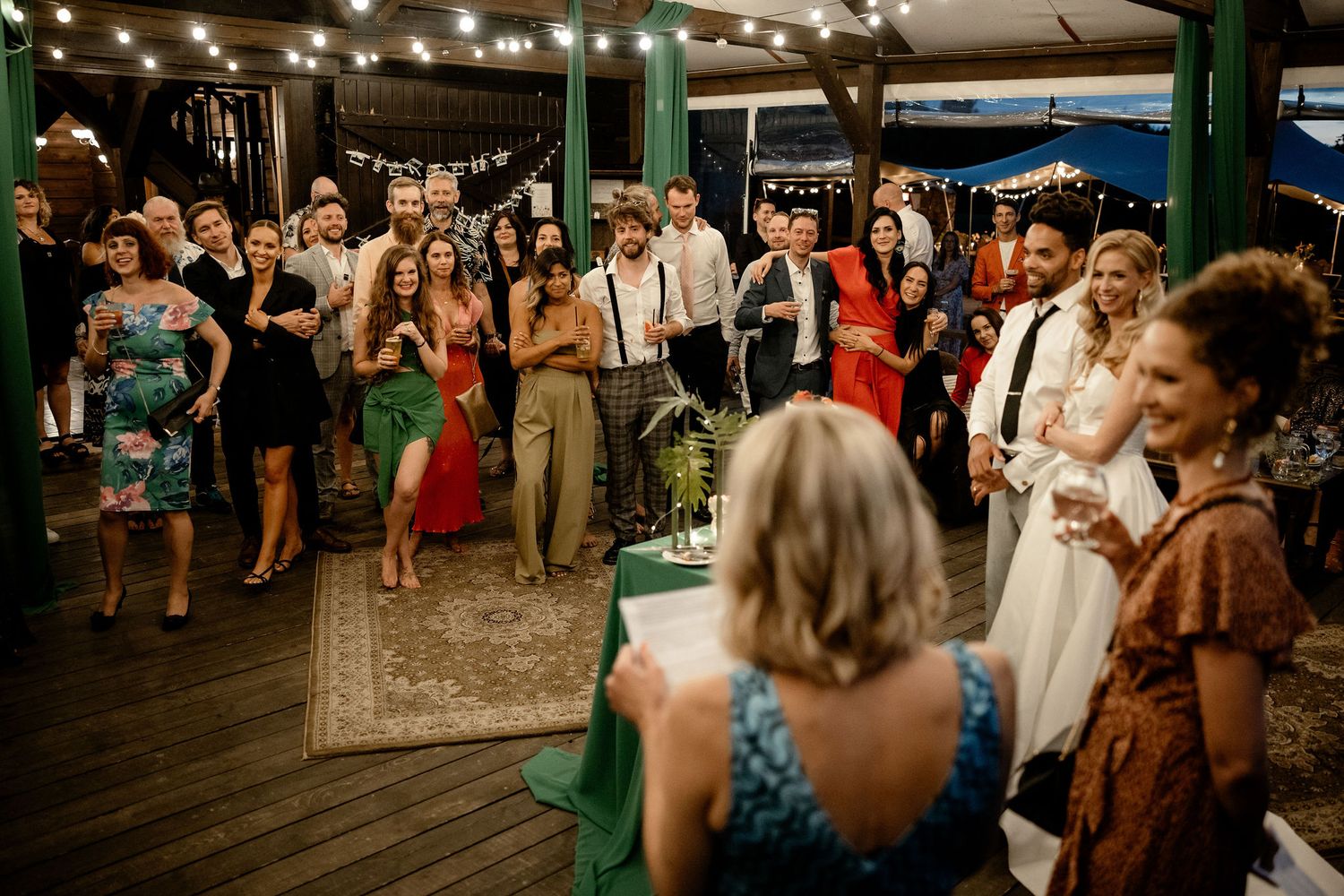 A lively wedding reception crowd dancing and celebrating on a covered outdoor patio with string lights overhead.