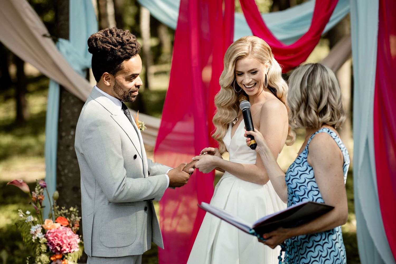 Intimate outdoor wedding ceremony with colorful fabric backdrop and floral arrangements in forest setting.