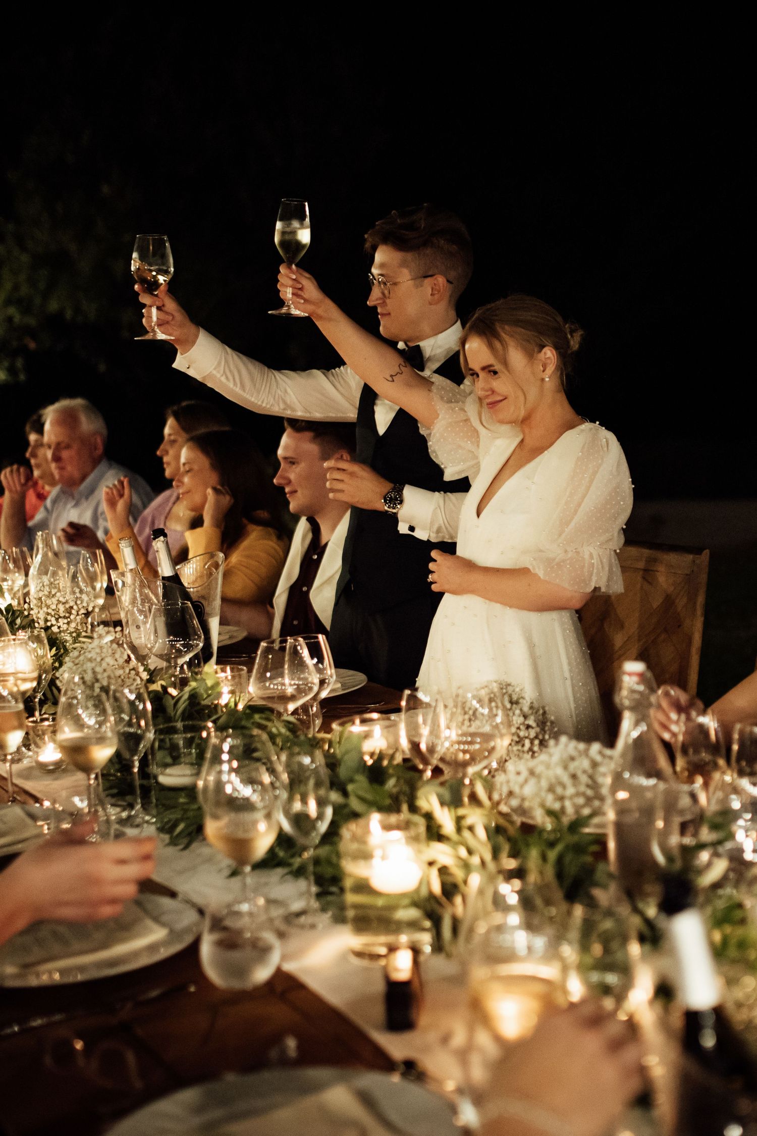 Celebratory toast at an elegant wedding reception with candlelit table decorated with greenery and white floral arrangements.