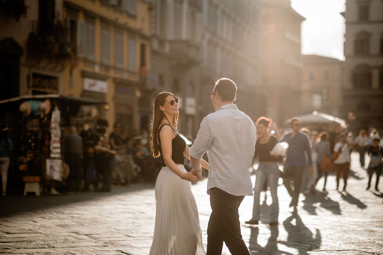 A couple in elegant attire strolls down a sunlit cobblestone street in an old European city during golden hour.