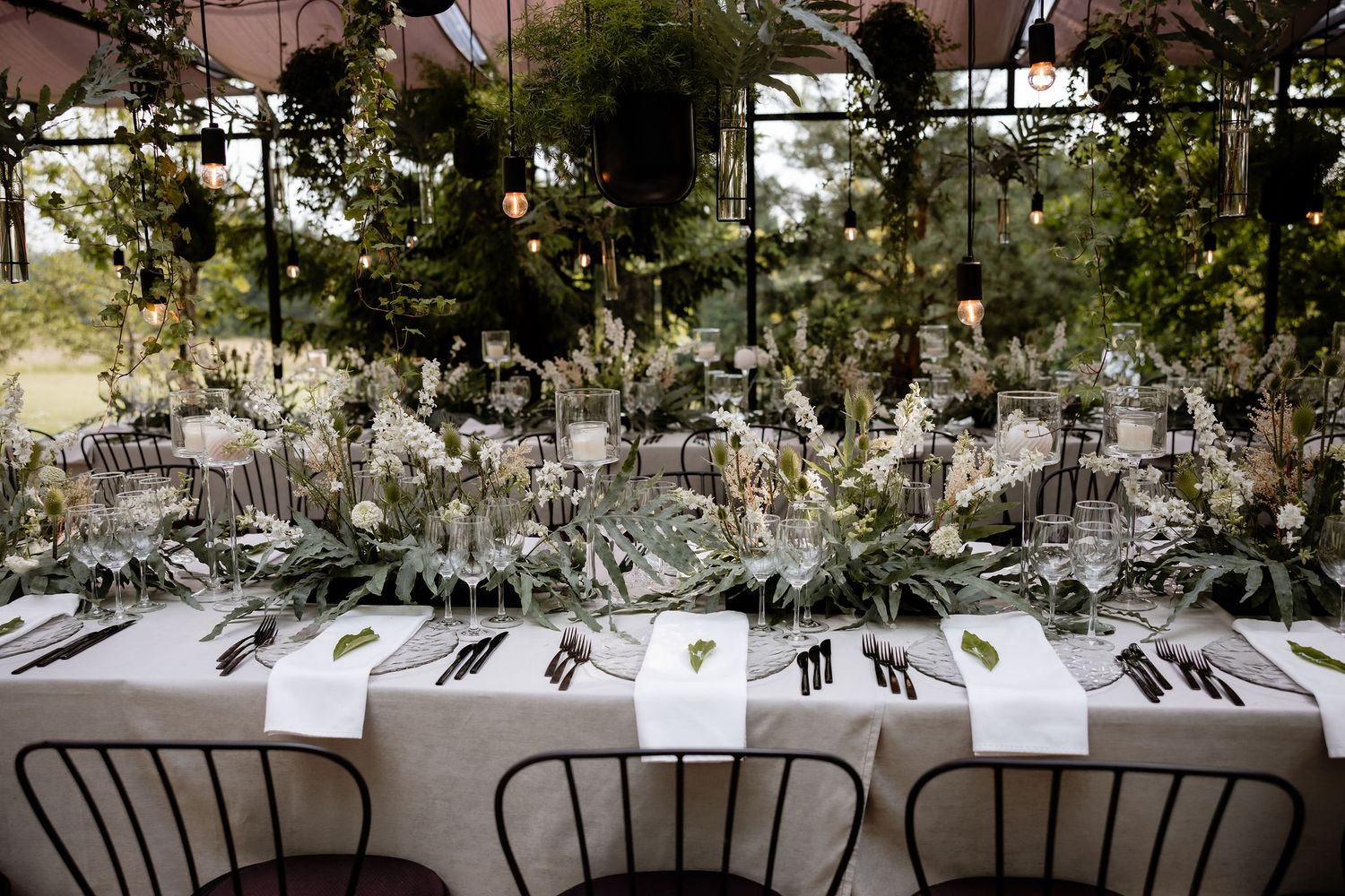 Long reception table decorated with greenery and elegant place settings in an indoor venue.