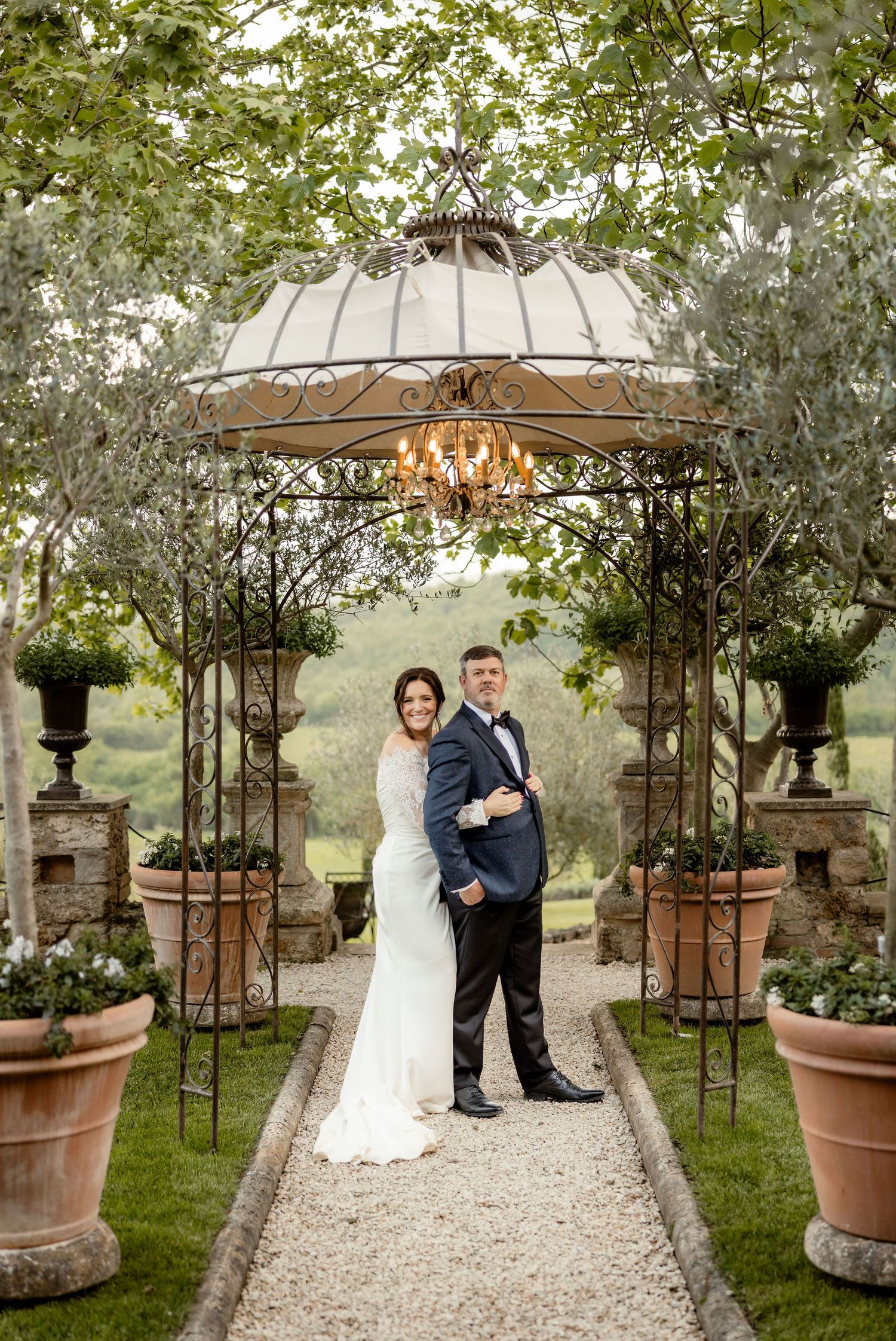 A romantic garden wedding ceremony setup with an ornate iron gazebo, gravel path and potted plants along the sides.