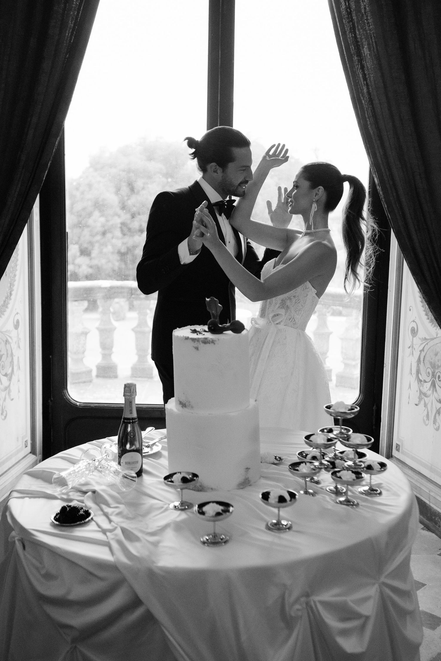 A black and white wedding photograph shows a couple playfully cutting their cake together at an elegant reception table.