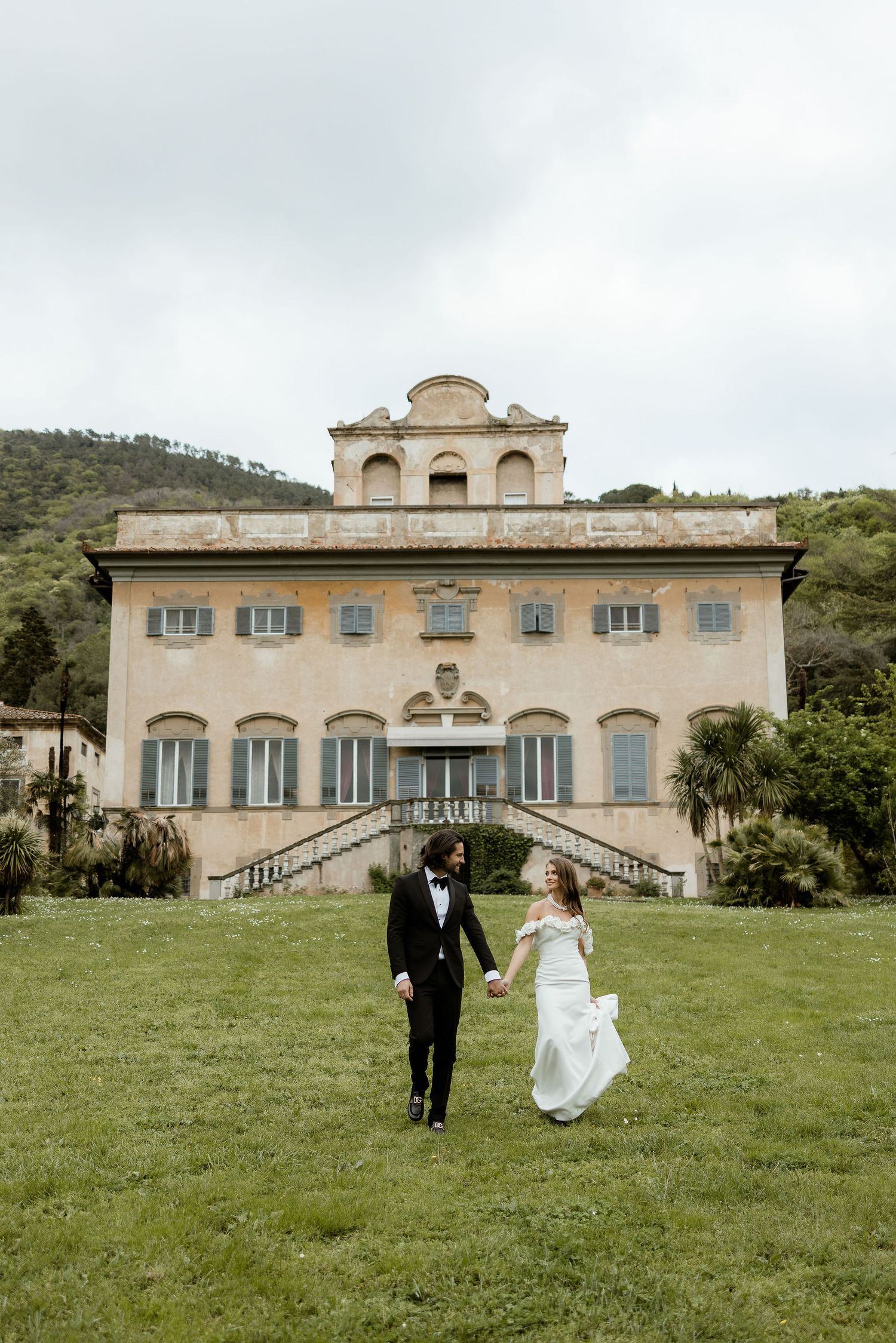 A couple in wedding attire walks hand in hand on grass in front of an elegant Italian villa with symmetrical architecture.