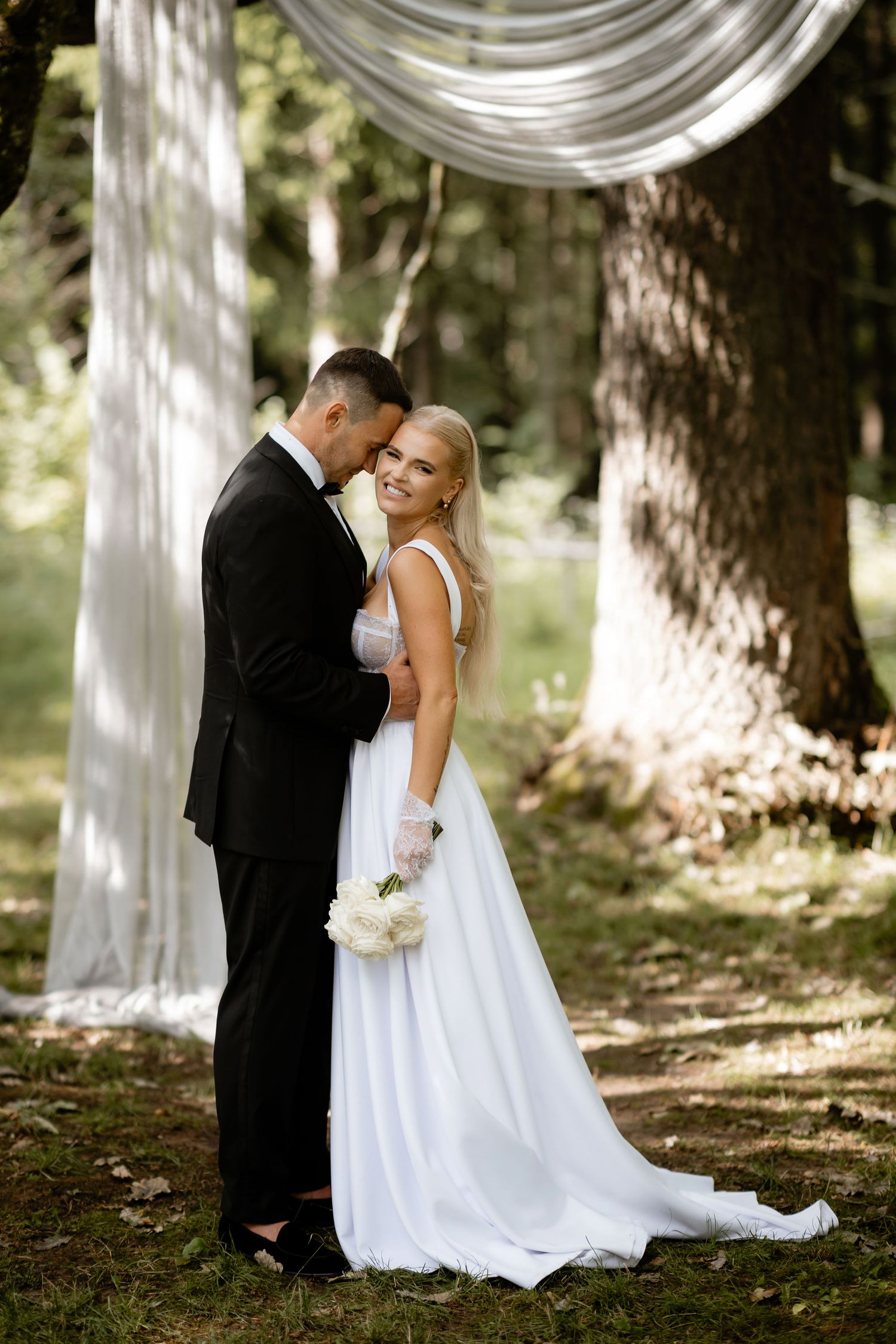 A romantic wedding moment captured under draped white fabric in a woodland setting.