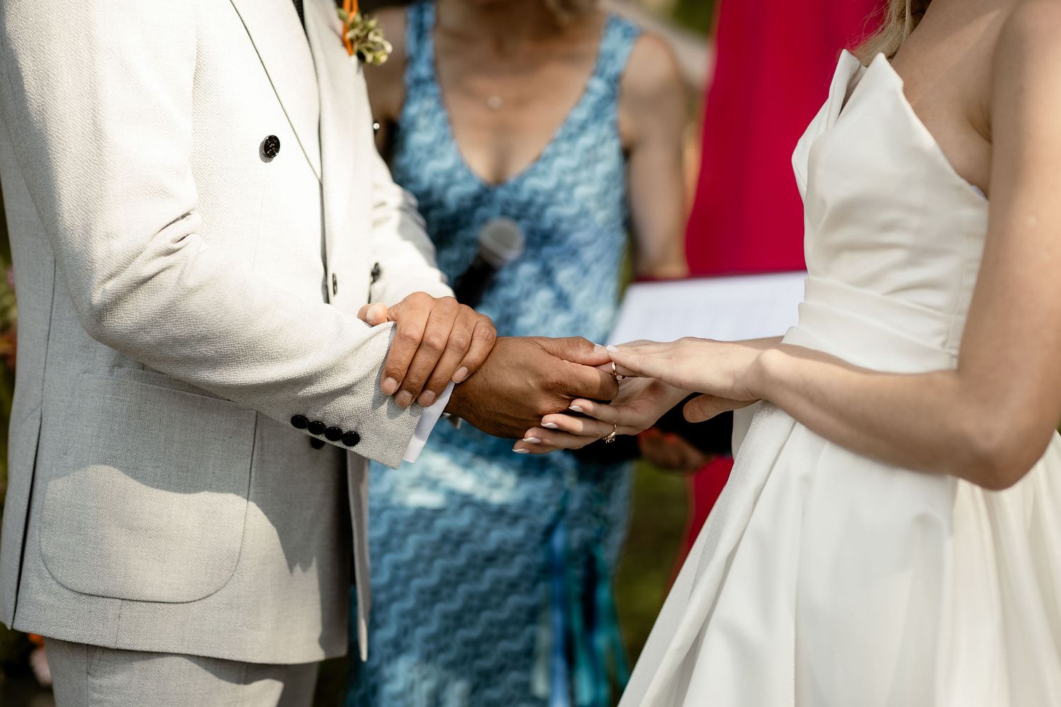 Close-up of hands during ring exchange at outdoor wedding ceremony with blue dress visible in background.