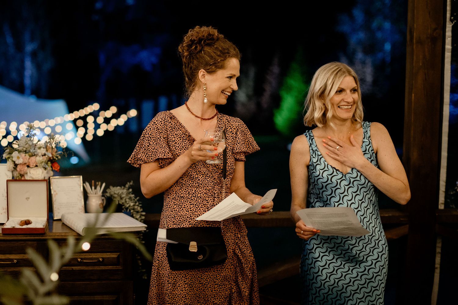 Two women reading from papers and laughing together during an evening outdoor event with string lights in background.