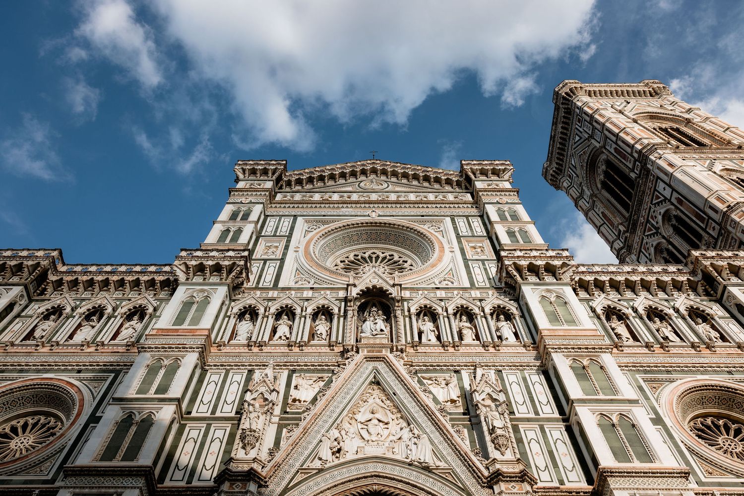 Looking up at the ornate marble facade of Florence Cathedral with its Gothic architecture and decorative details against a blue sky.