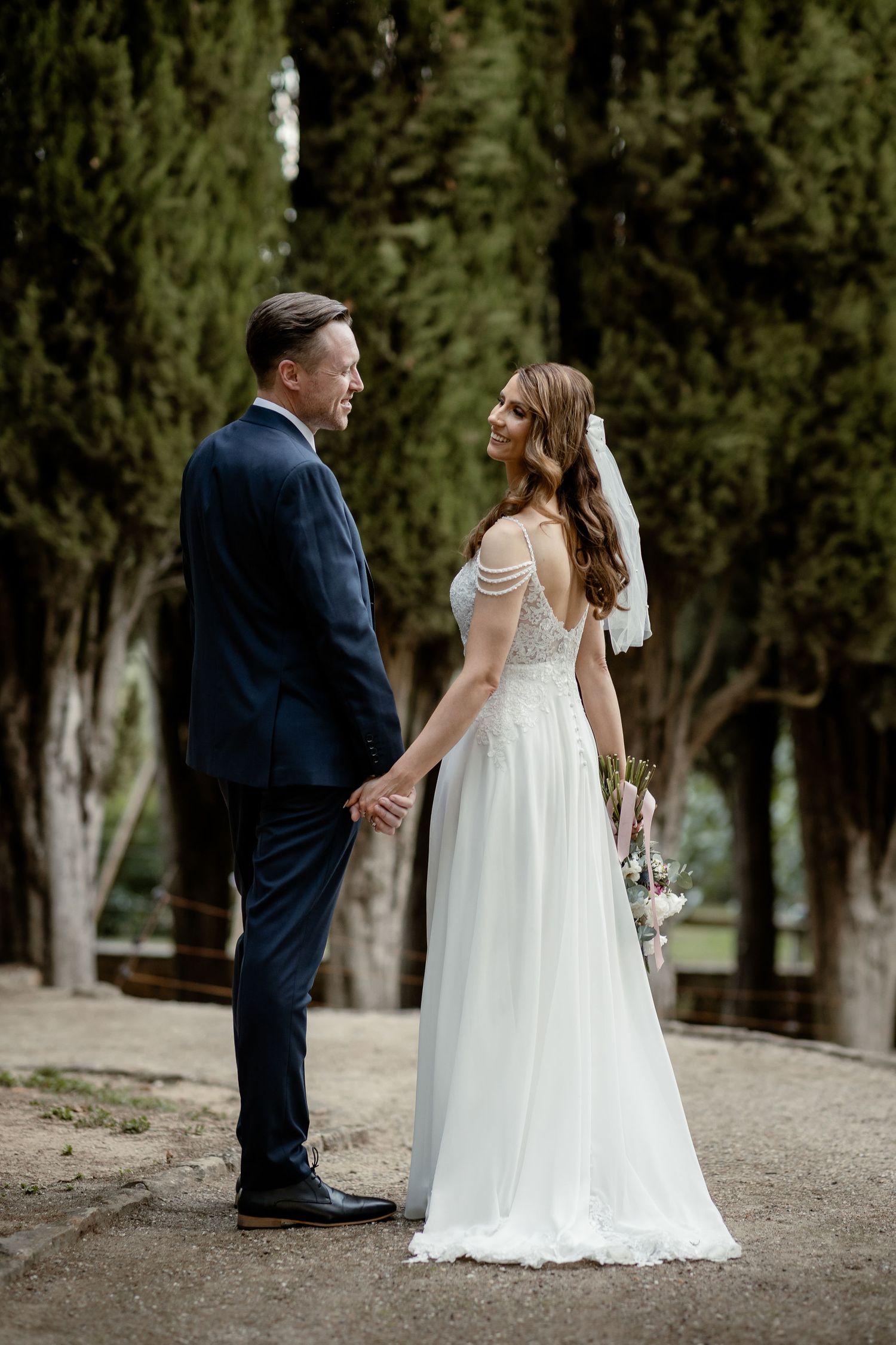 Newlyweds in elegant attire stand together on a tree-lined path during their wedding photoshoot.