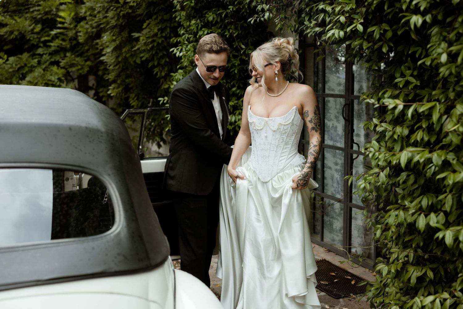 A bride in a flowing white wedding dress poses among green foliage and trees in an outdoor setting.