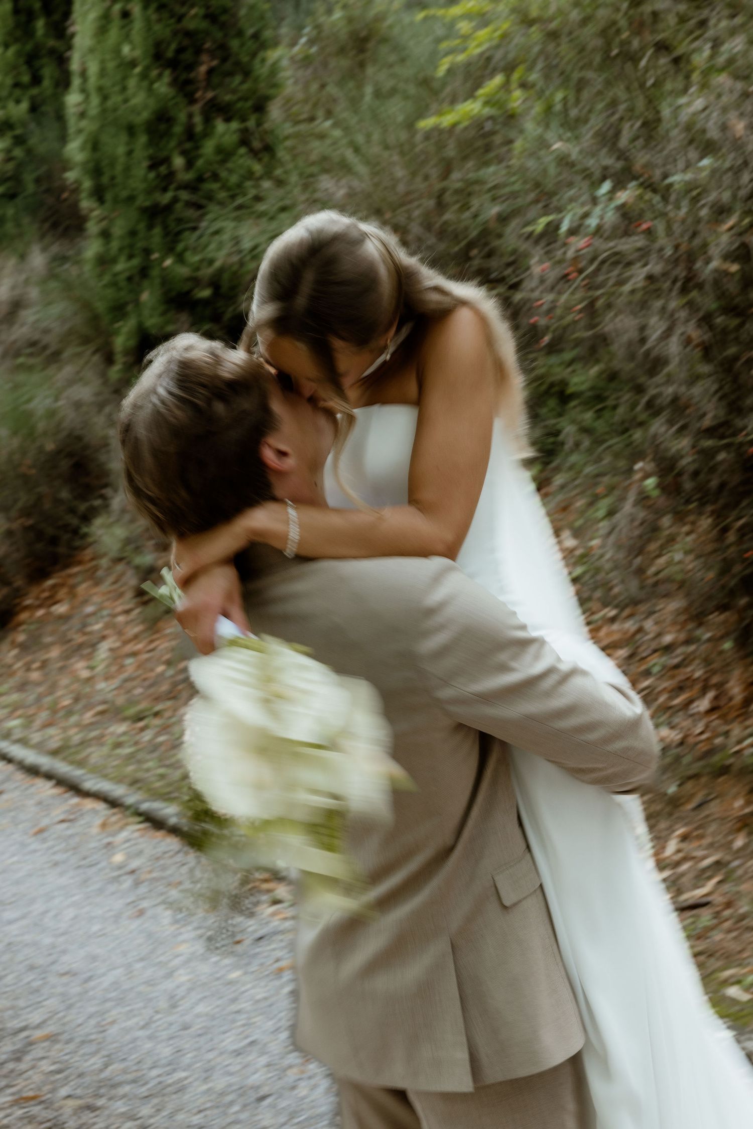 Newlyweds share an intimate moment outdoors with flowing white veil and beige suit.