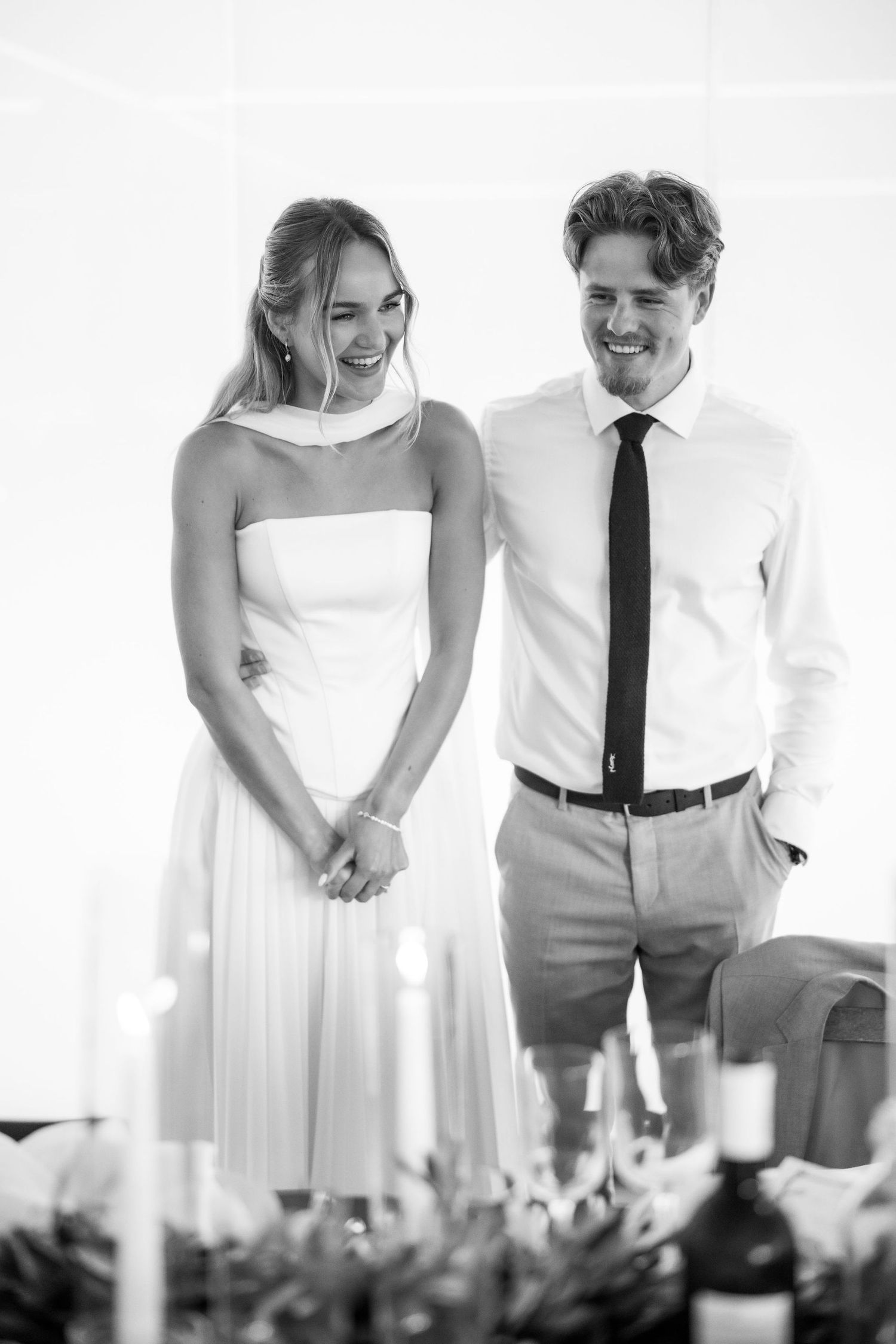 A black and white candid wedding reception moment showing a couple in formal attire laughing together at their table.