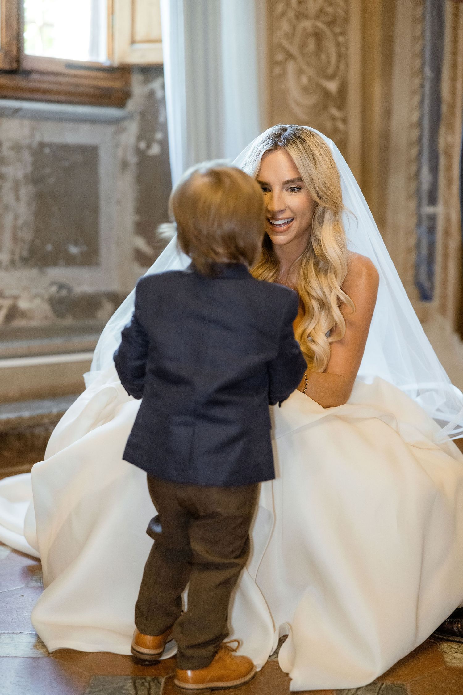A bride in a white wedding dress kneels down to interact with a small boy in a dark suit at an elegant venue.