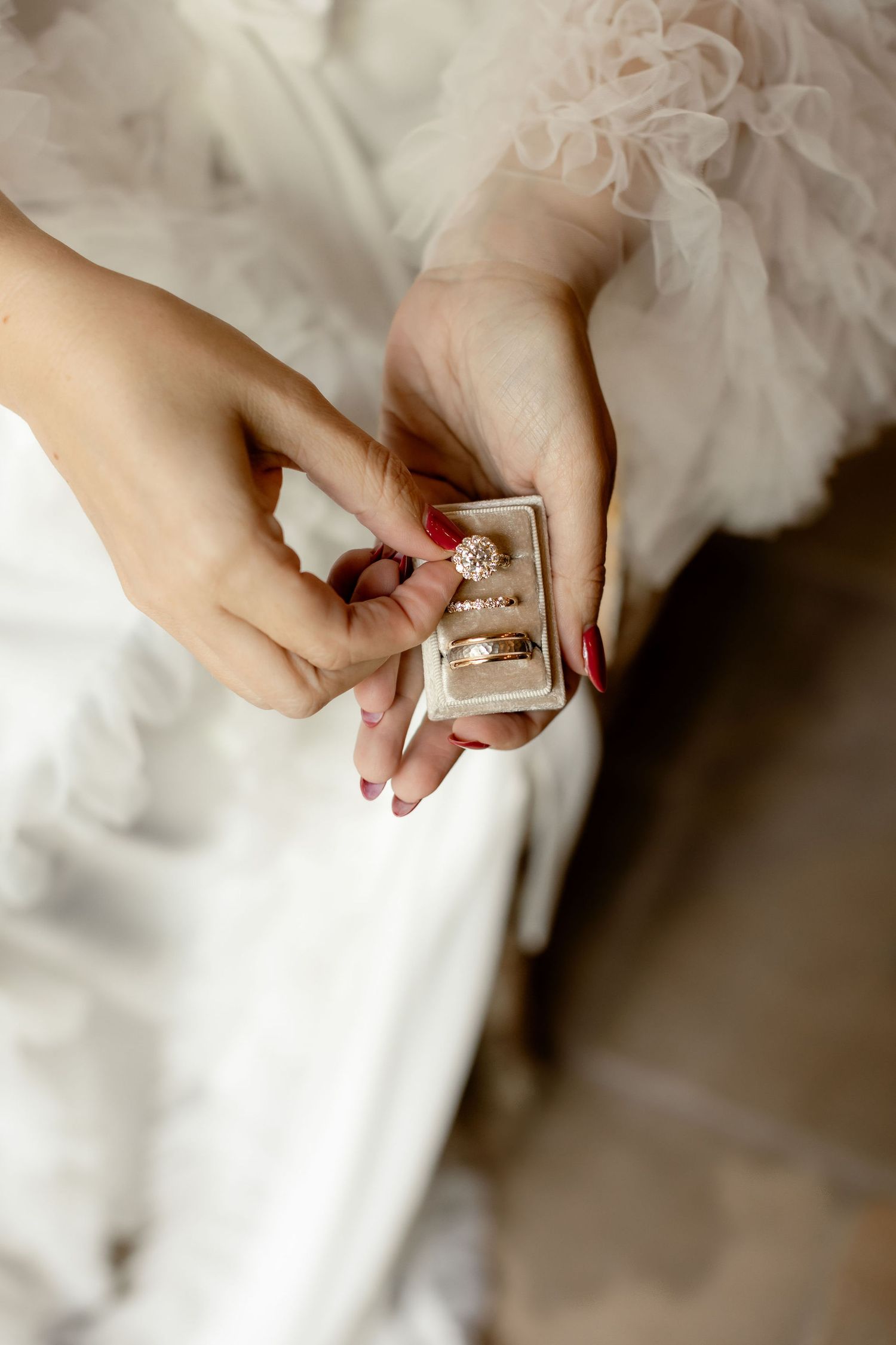 Hands holding an elegant engagement ring box with red manicured nails against white wedding dress fabric.