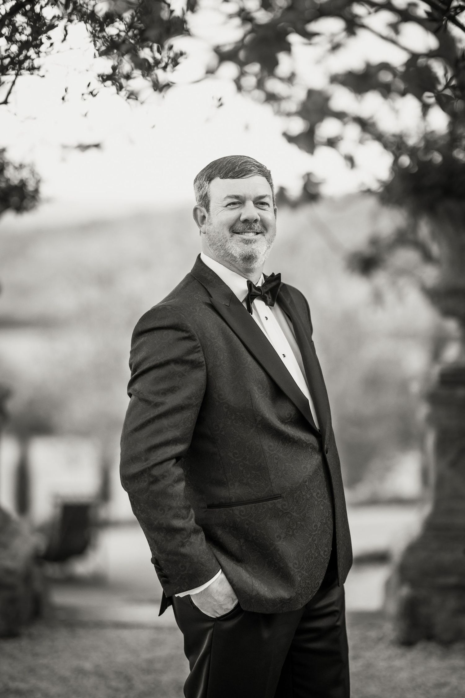 Black and white portrait of a groom in a formal tuxedo standing outdoors with a confident pose.