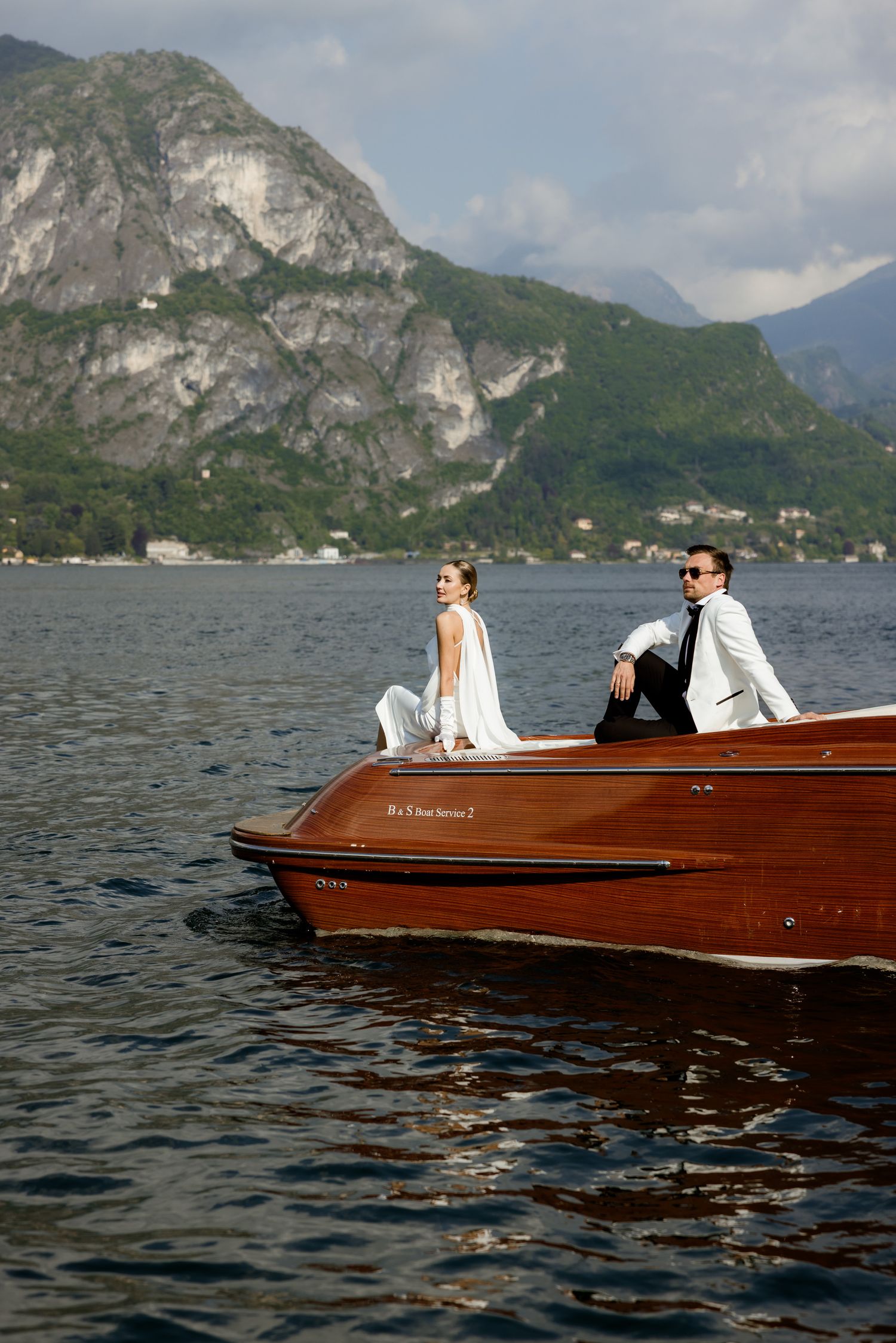 Elegant couple in white wedding attire sitting on a classic wooden boat on a serene lake with mountain backdrop.