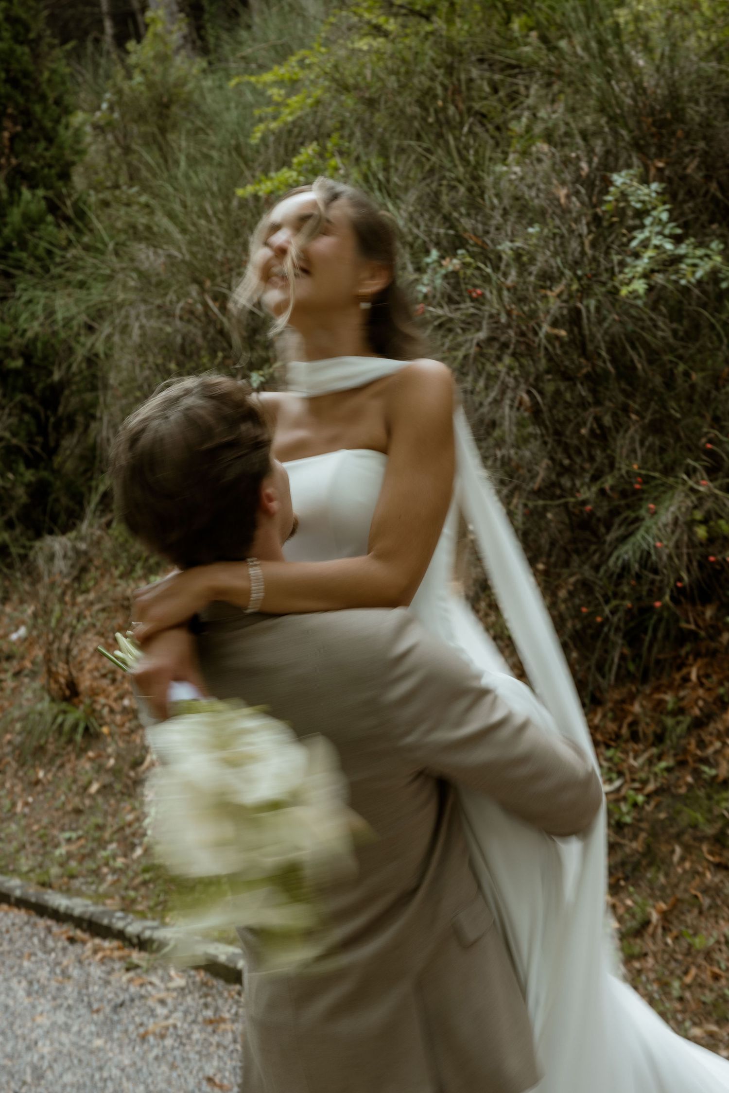 A couple in wedding attire spins joyfully in an embrace on a wooded path during their celebration.