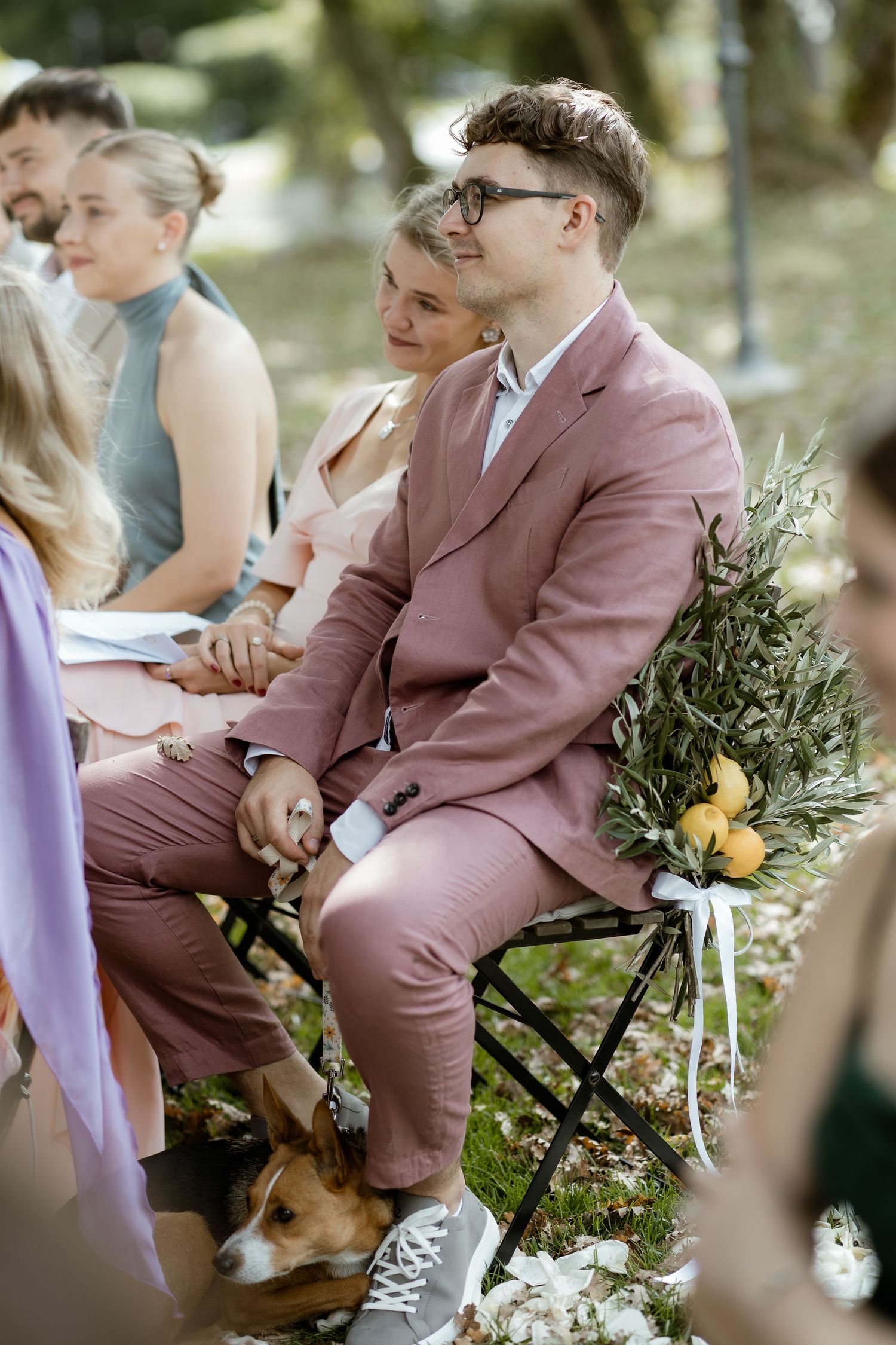 Wedding guests in mauve suits sit outdoors on folding chairs during an outdoor ceremony with citrus and greenery decor.