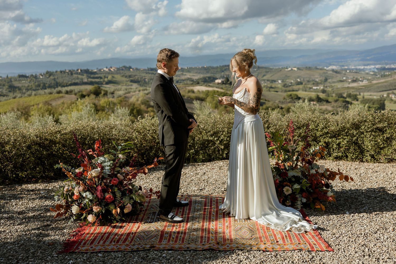 A couple stands together on a scenic hilltop overlooking a vast landscape with a red rug and floral arrangements.