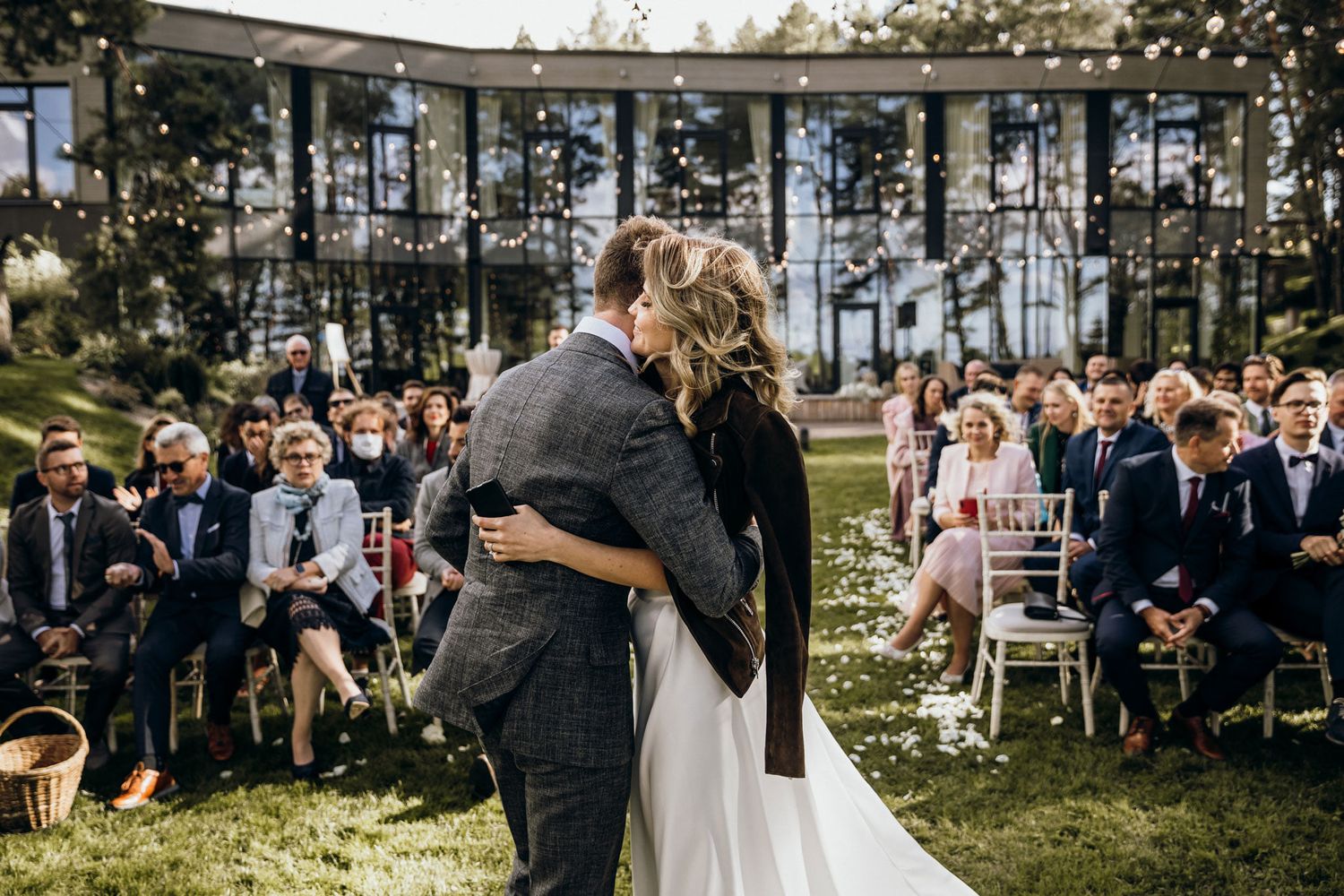 Couple exchanges vows during outdoor wedding ceremony surrounded by guests seated on white chairs under tall trees.