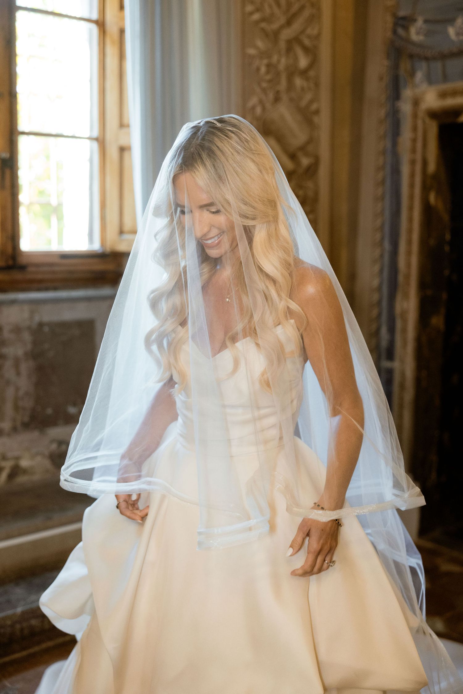 A bride wearing a classic white ballgown and veil smiles joyfully in an ornate room with natural lighting.
