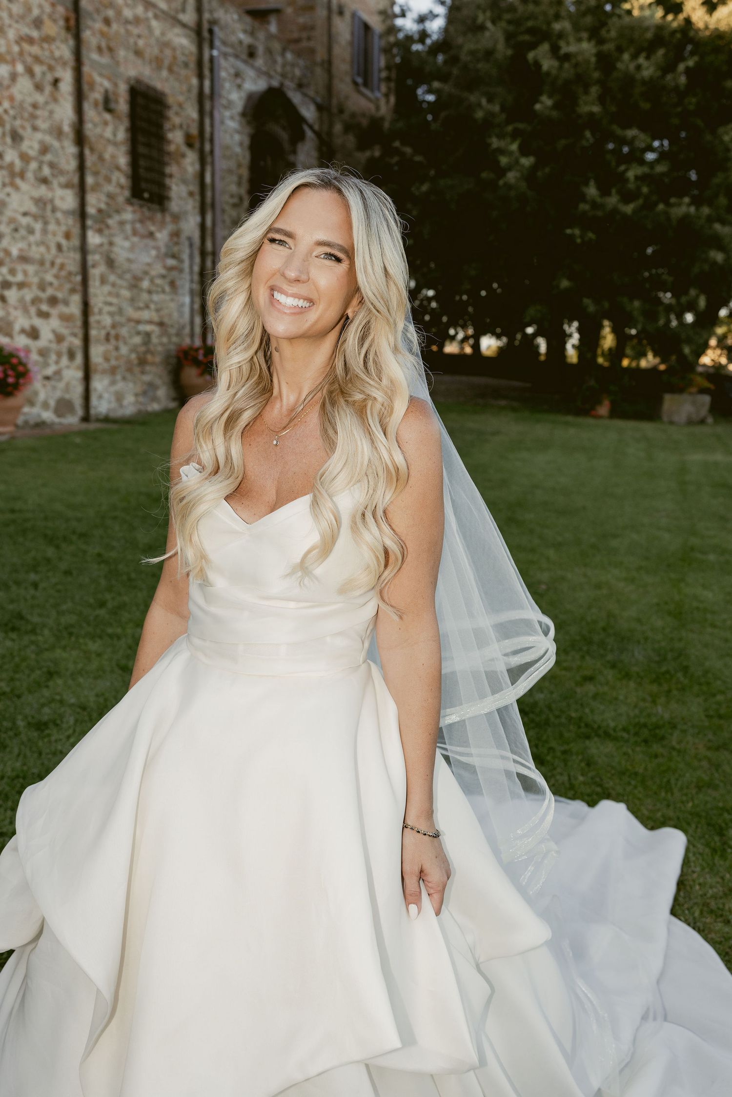 A bride in a flowing white wedding dress twirls joyfully on a grassy hillside at sunset.
