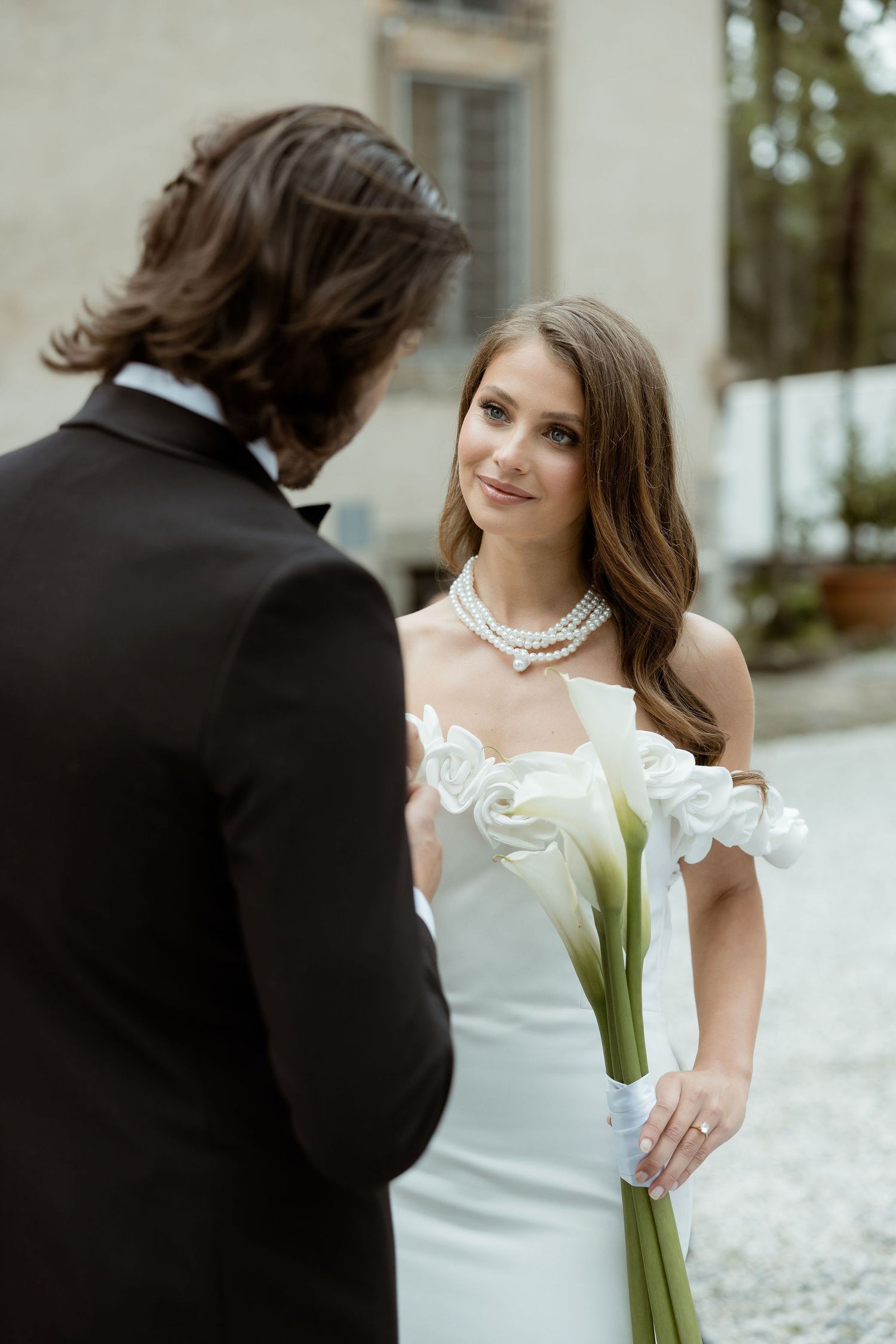 A bride in an off-shoulder white gown holds a bouquet of calla lilies while looking at her groom in a dark suit.