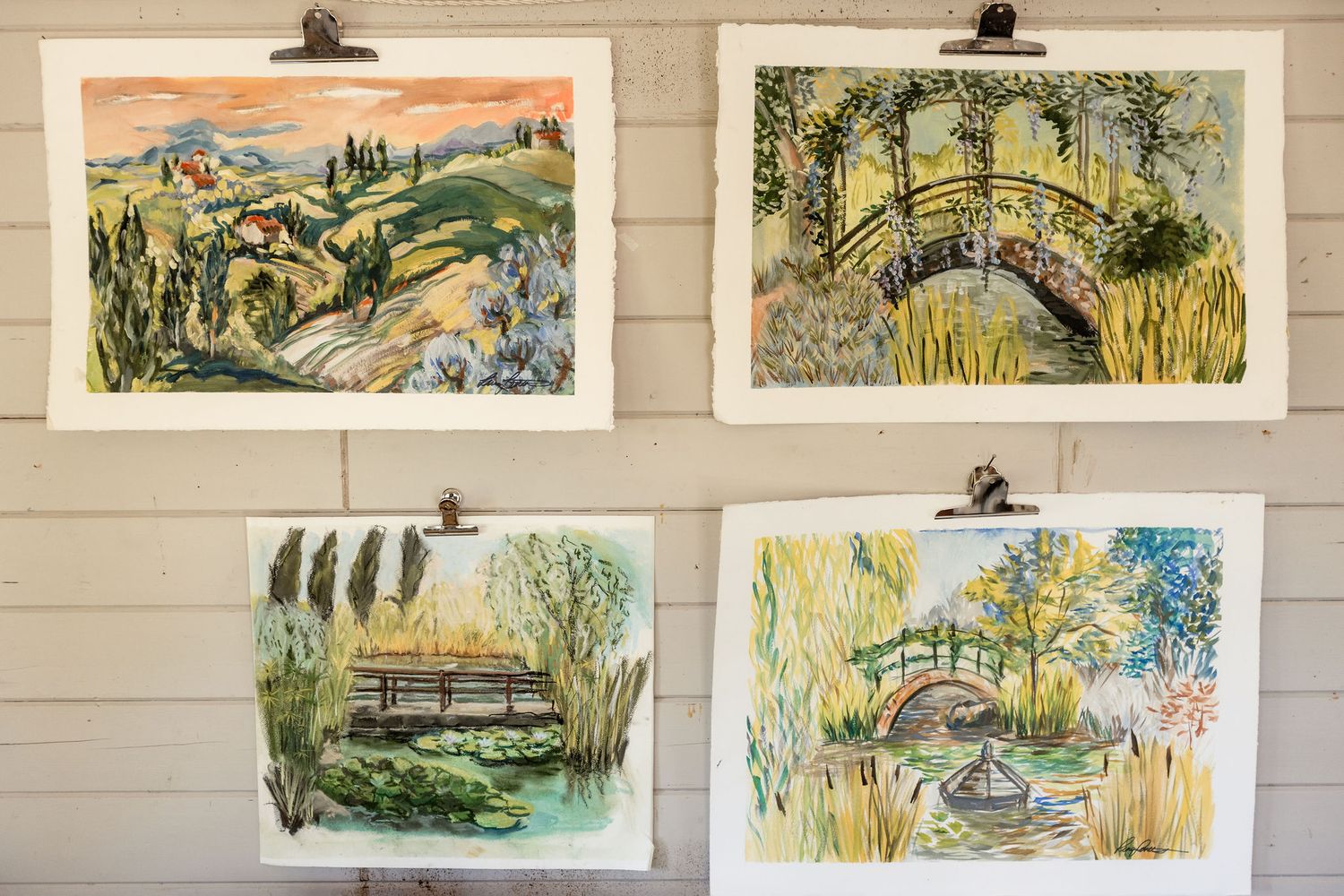 A wall gallery displays multiple framed watercolor landscape artworks featuring rural scenes and natural settings.