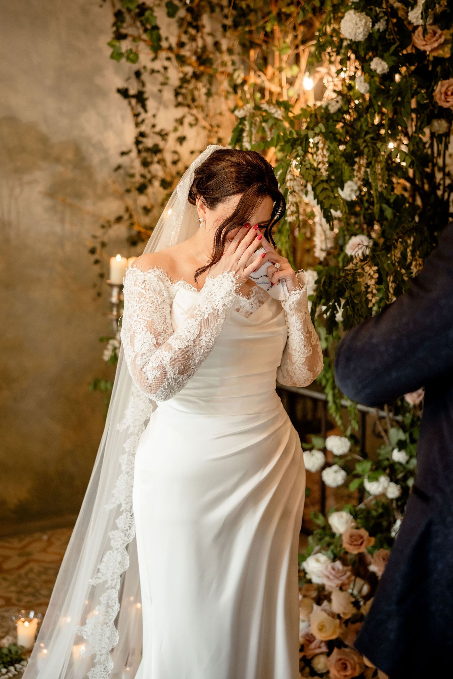 A bride in a long-sleeved lace wedding gown wipes tears during an emotional moment beside a floral arch.