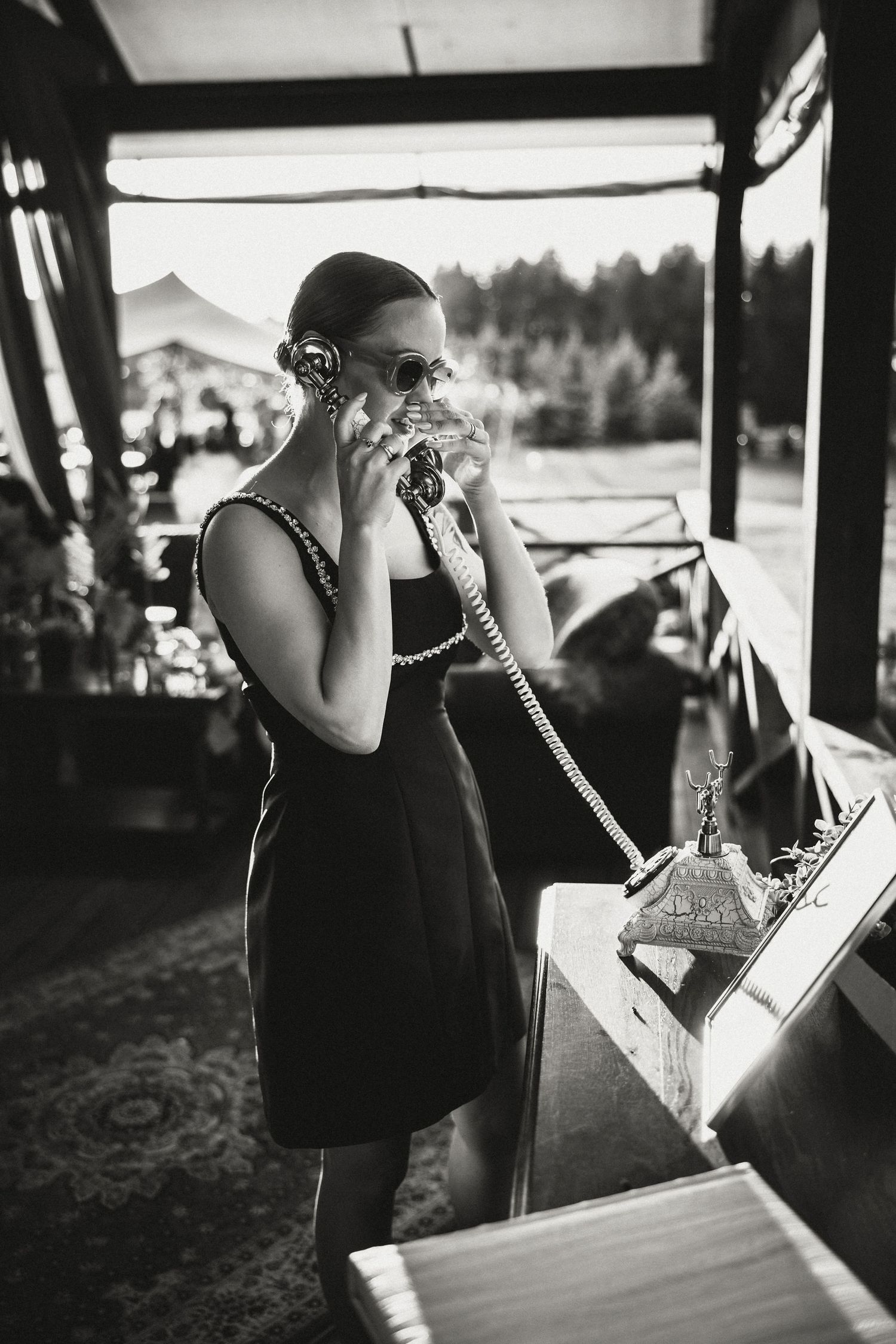 Person in black dress making call on vintage telephone at outdoor event.