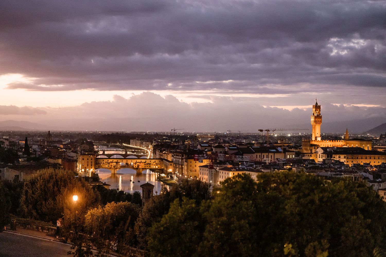 Dramatic sunset view of Florence cityscape with illuminated landmarks and moody storm clouds overhead.