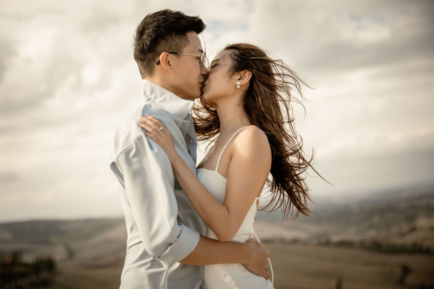 Intimate portrait of couple sharing kiss against dramatic Tuscan countryside.