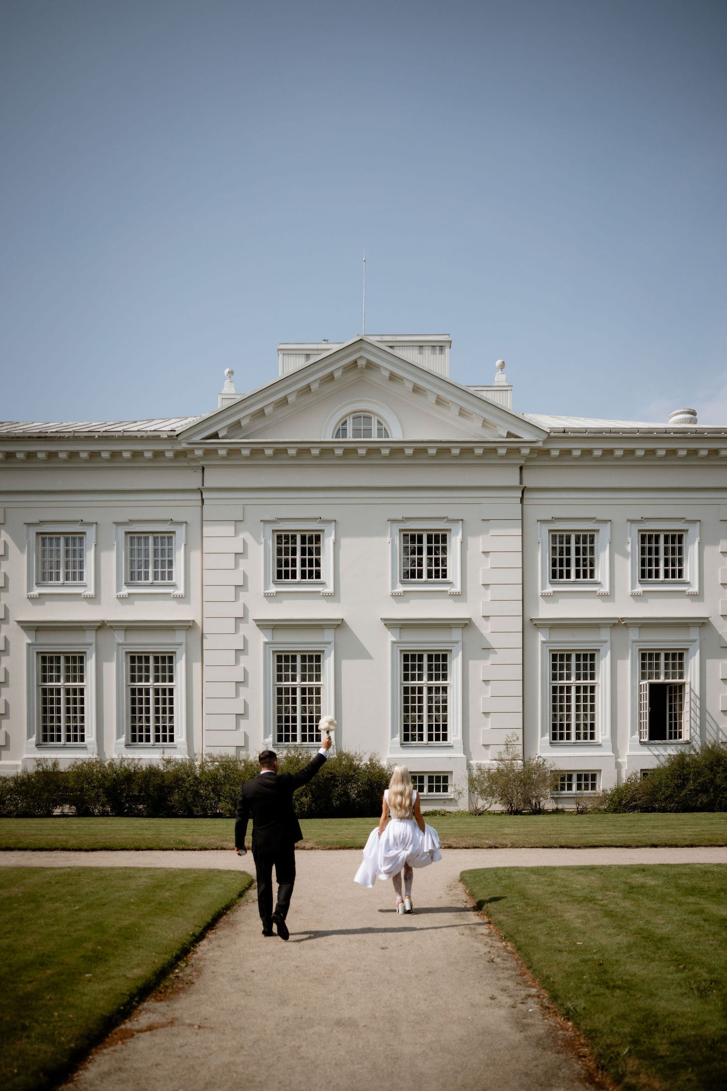 Two people walking towards an elegant white mansion with classical architecture and manicured lawns.