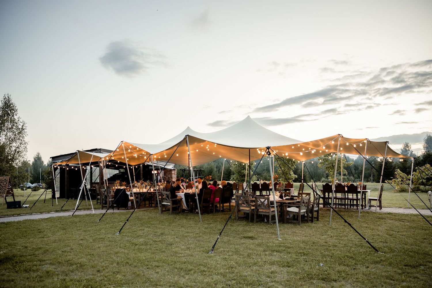 Outdoor event setup with string lights and tent canopy at sunset in a grassy field surrounded by trees.