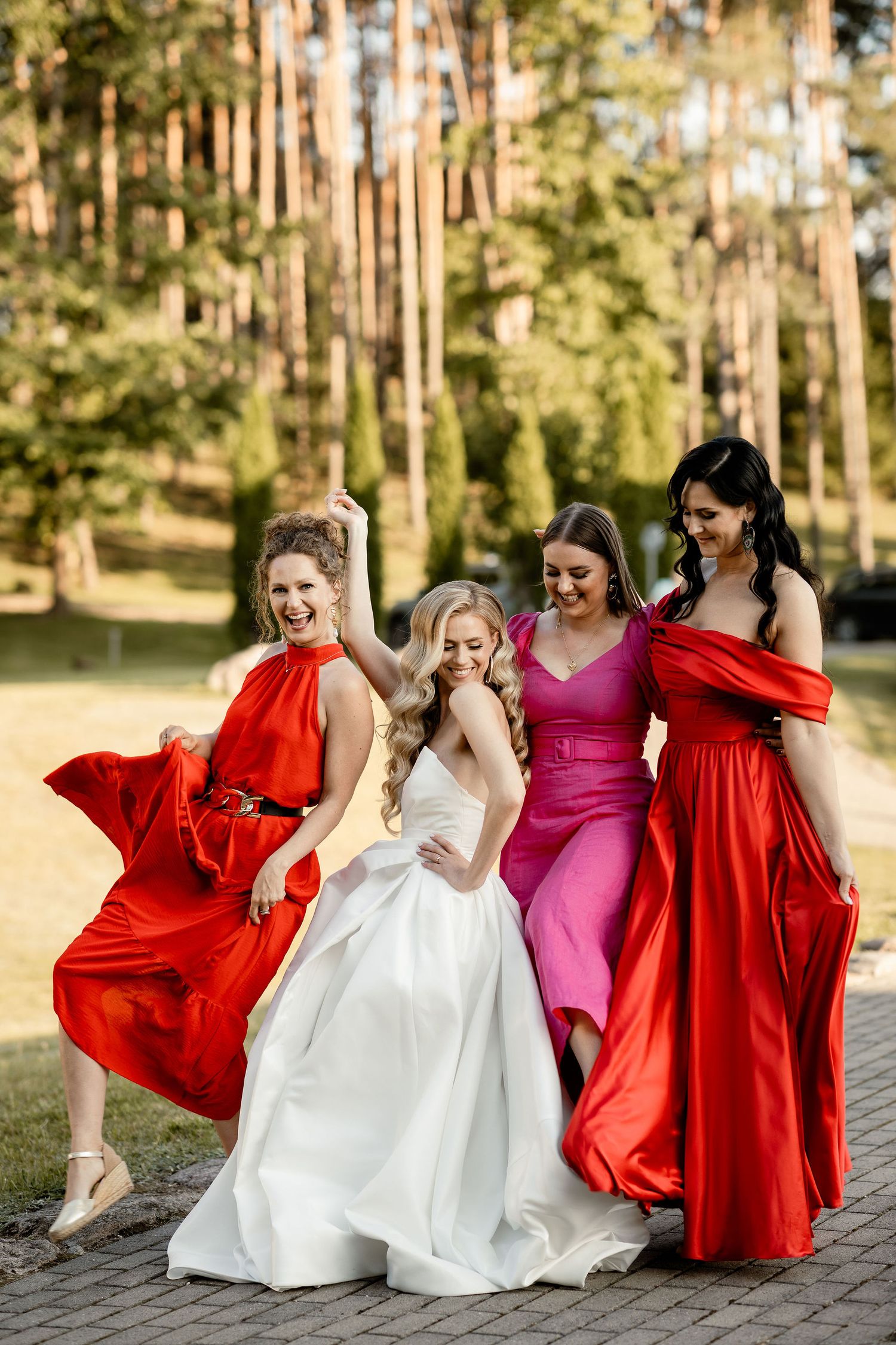 Wedding party in vibrant red dresses and white gown pose playfully together on path with forest backdrop.