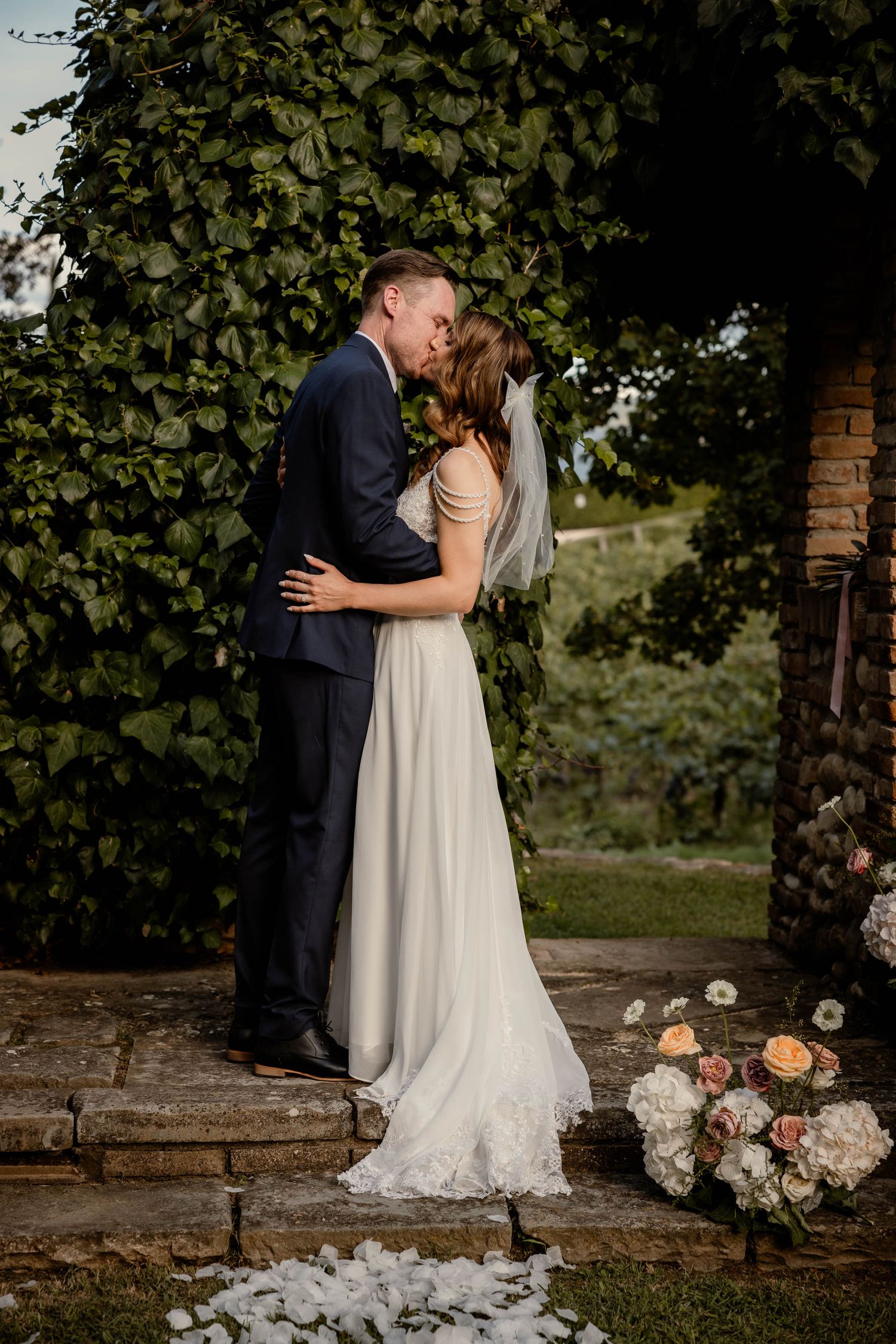 Romantic wedding couple embraces on garden steps with floral decor and ivy backdrop.