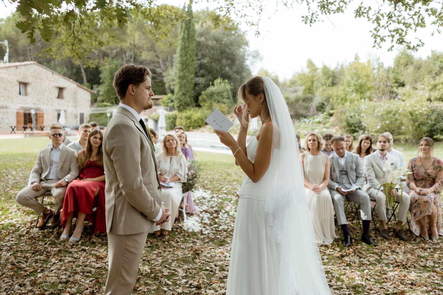 A romantic outdoor wedding ceremony takes place in a lush garden setting with guests watching the couple exchange vows.