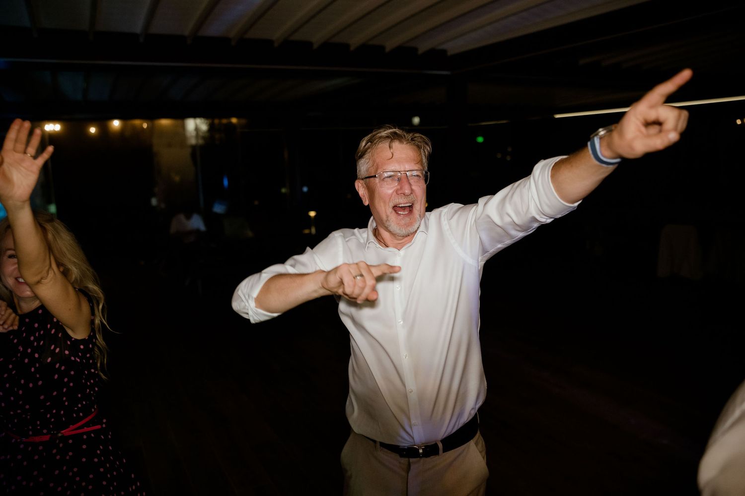 A person in a white dress shirt dances enthusiastically with arms outstretched at a nighttime event.