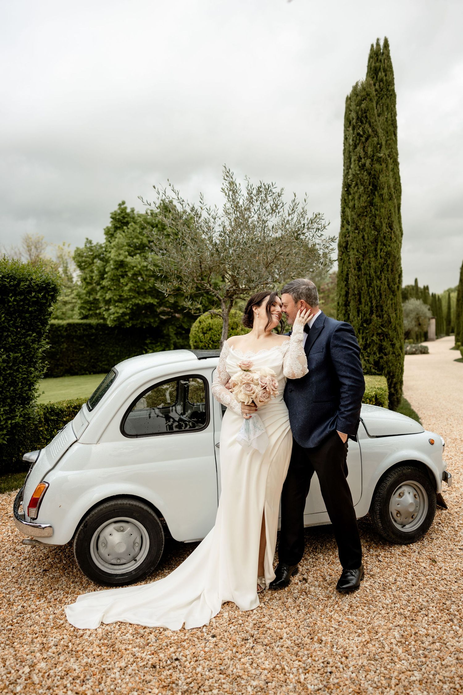 A couple shares a romantic moment next to a vintage Fiat 500 on a gravel driveway with cypress trees in the background.