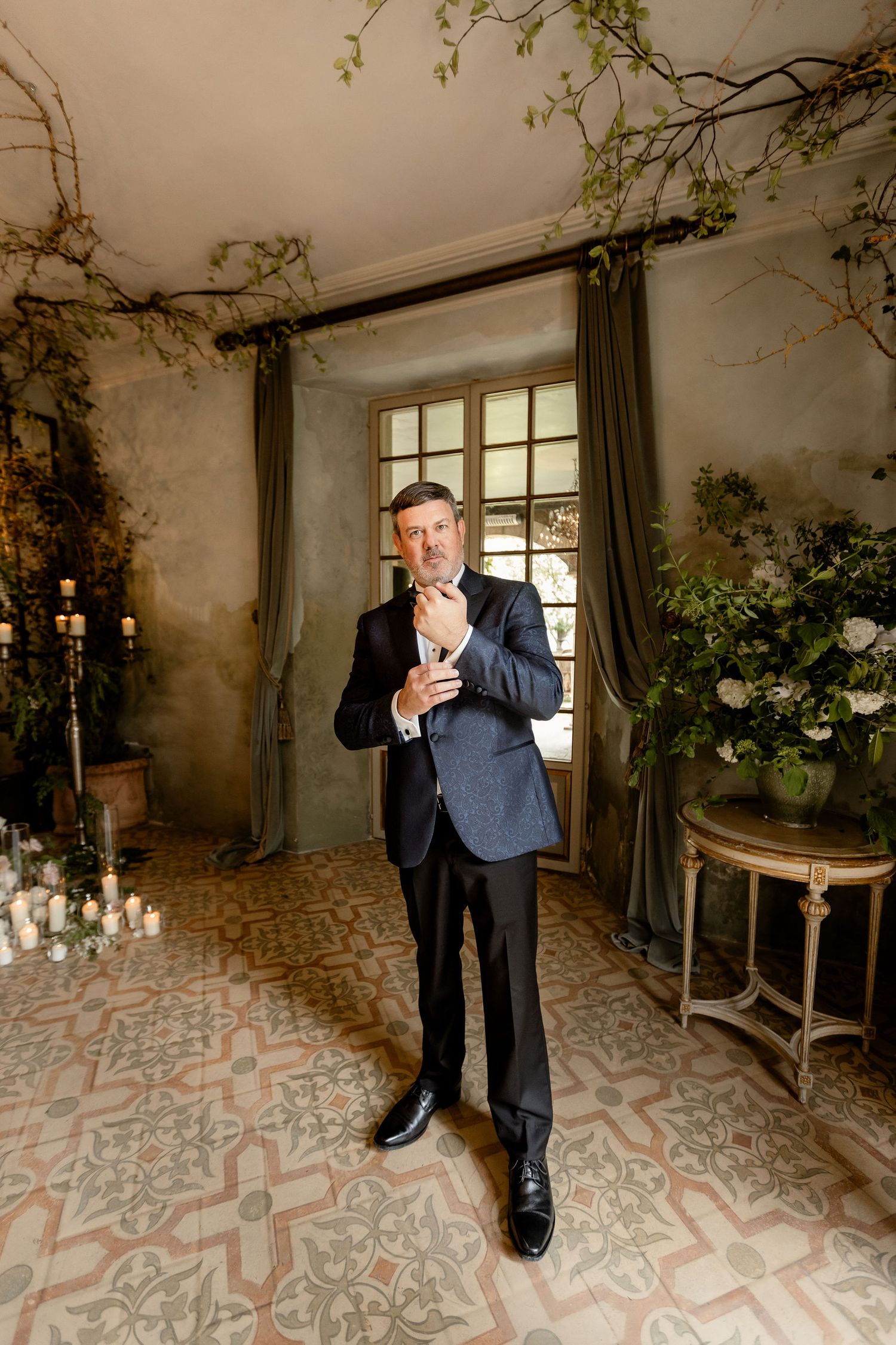 A person in a dark suit stands in an elegant room with patterned floors, floral arrangements, and draped windows.