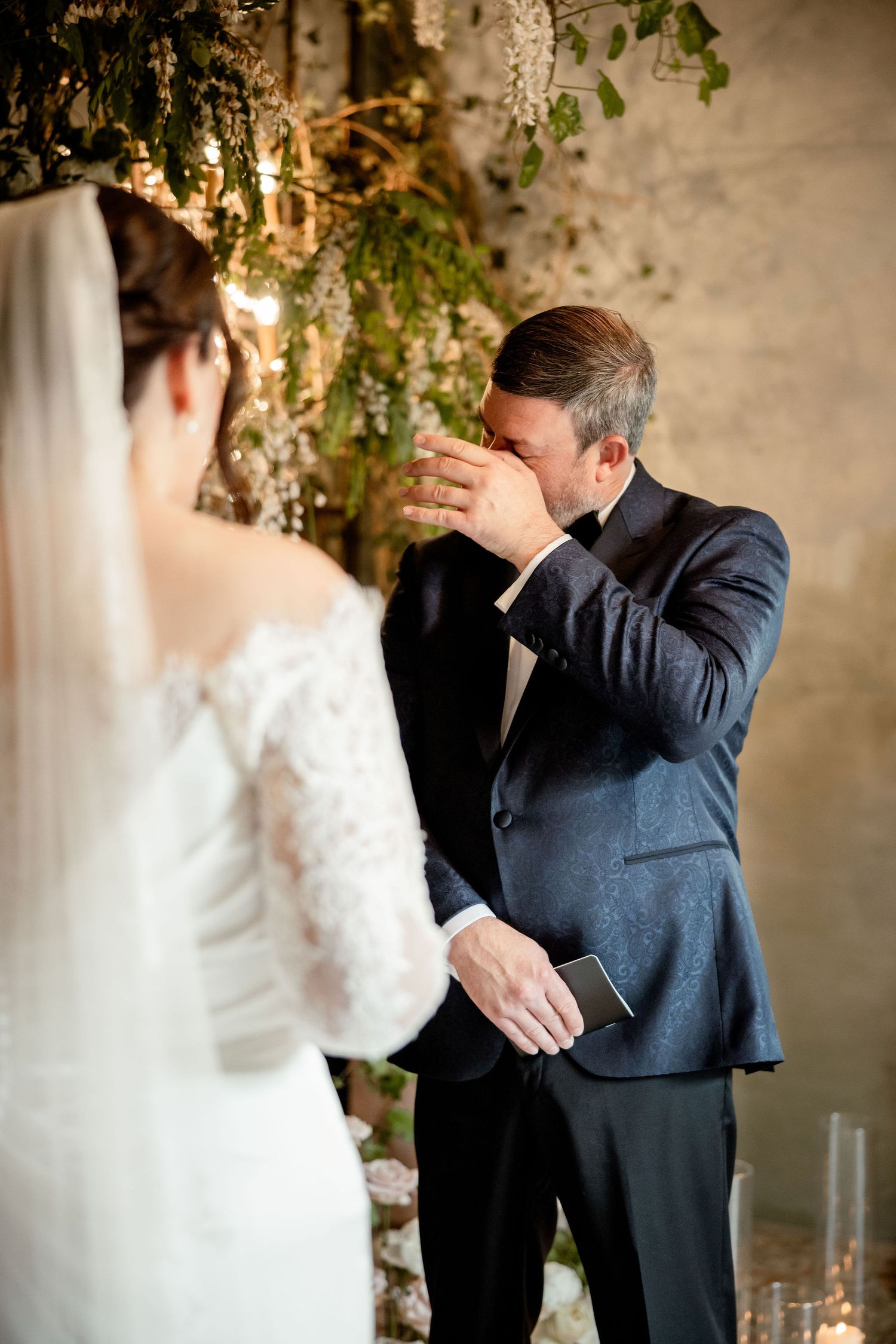 An emotional groom covers his face during a first look wedding moment as his bride stands nearby in white.