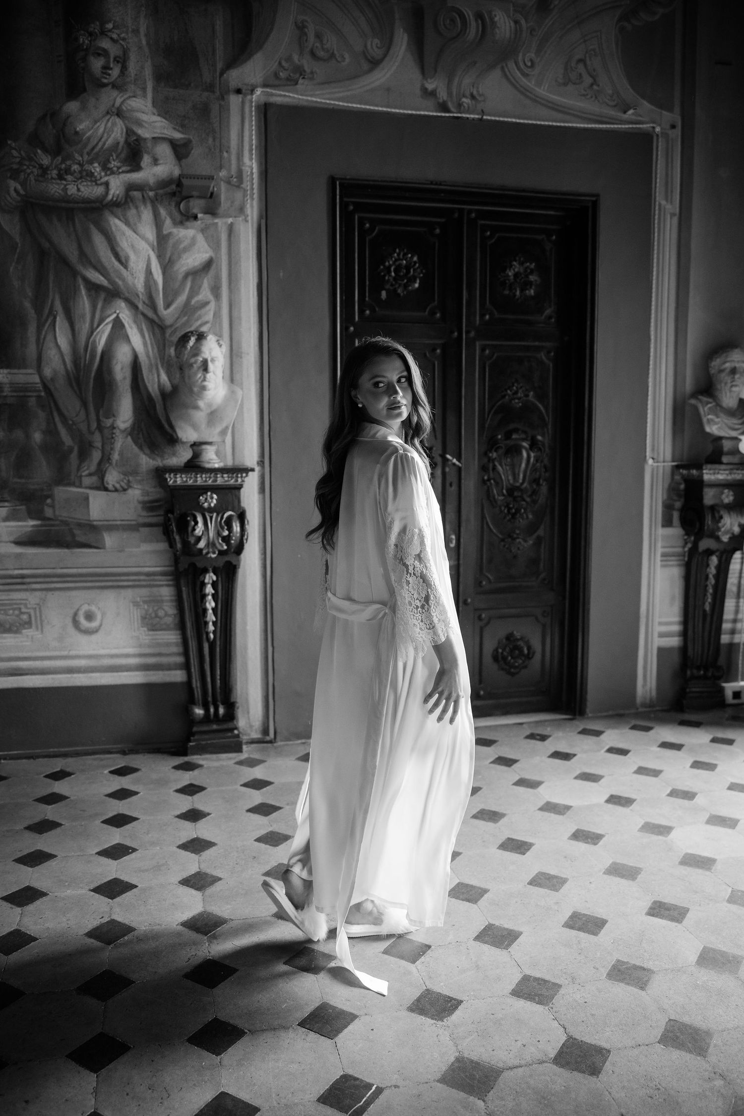 A figure in a flowing white dress stands in an ornate historic hallway with patterned floor tiles and decorative wall sculptures.
