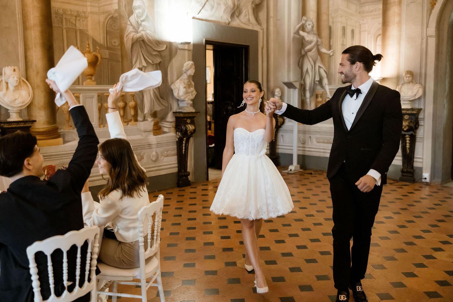 A couple celebrates in an elegant ballroom with ornate decor and tiled floors as guests look on with excitement.